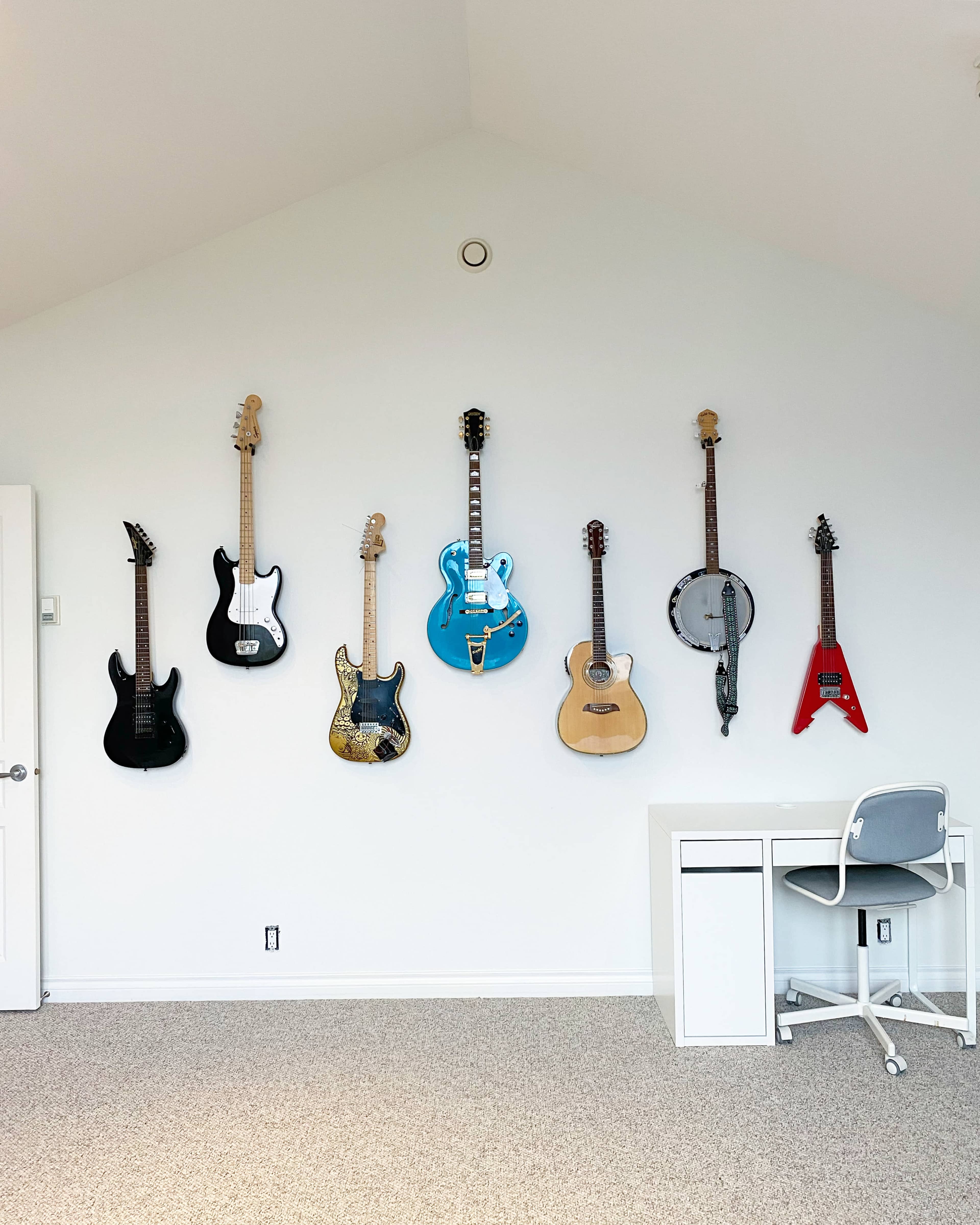 A variety of musical instruments, including electric guitars, an acoustic guitar, a banjo, and a red guitar, are displayed on a white wall above a small white desk and chair in a carpeted room.