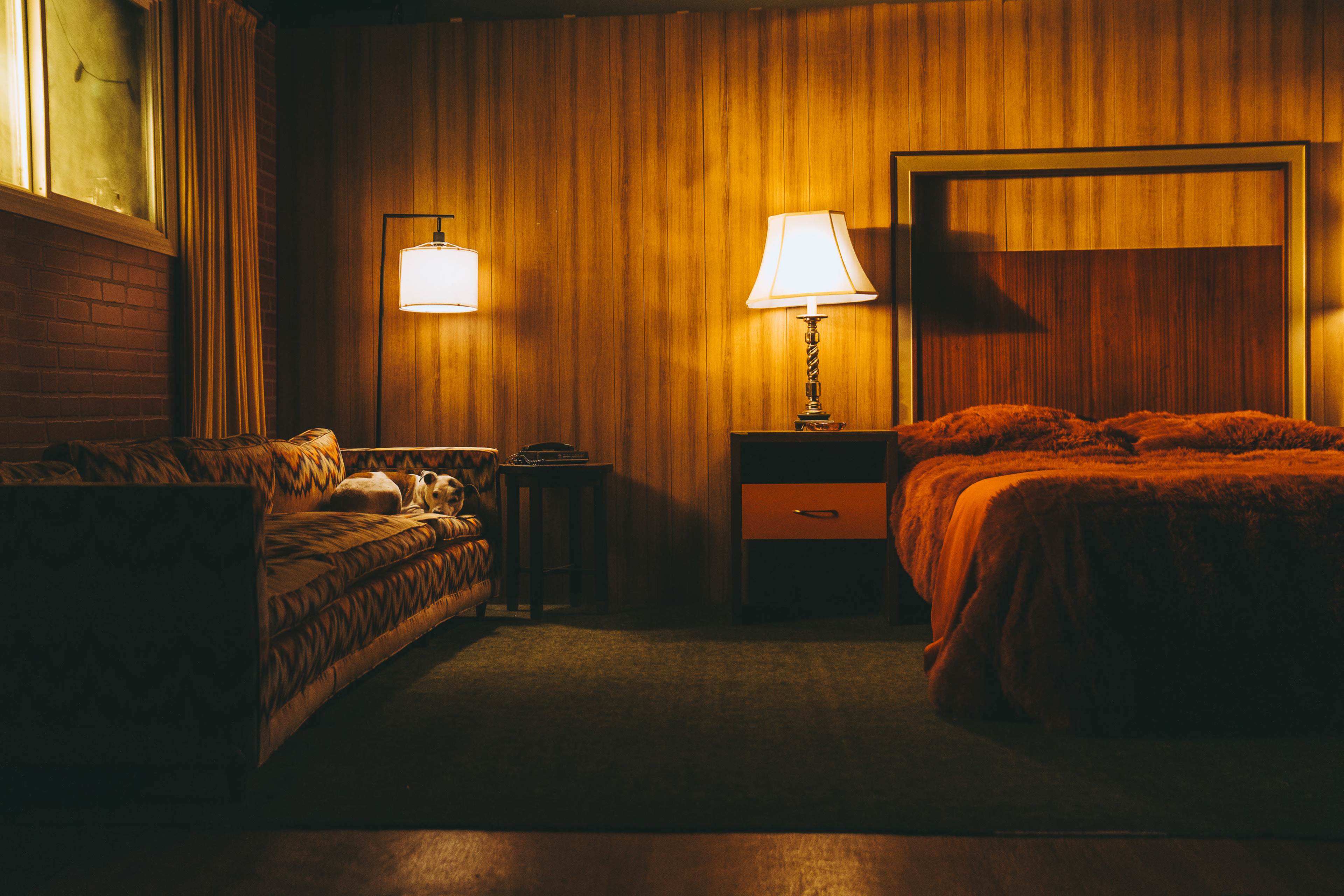 A cozy room features a bed with orange bedding, a patterned couch, and soft lighting from a lamp beside the bed.