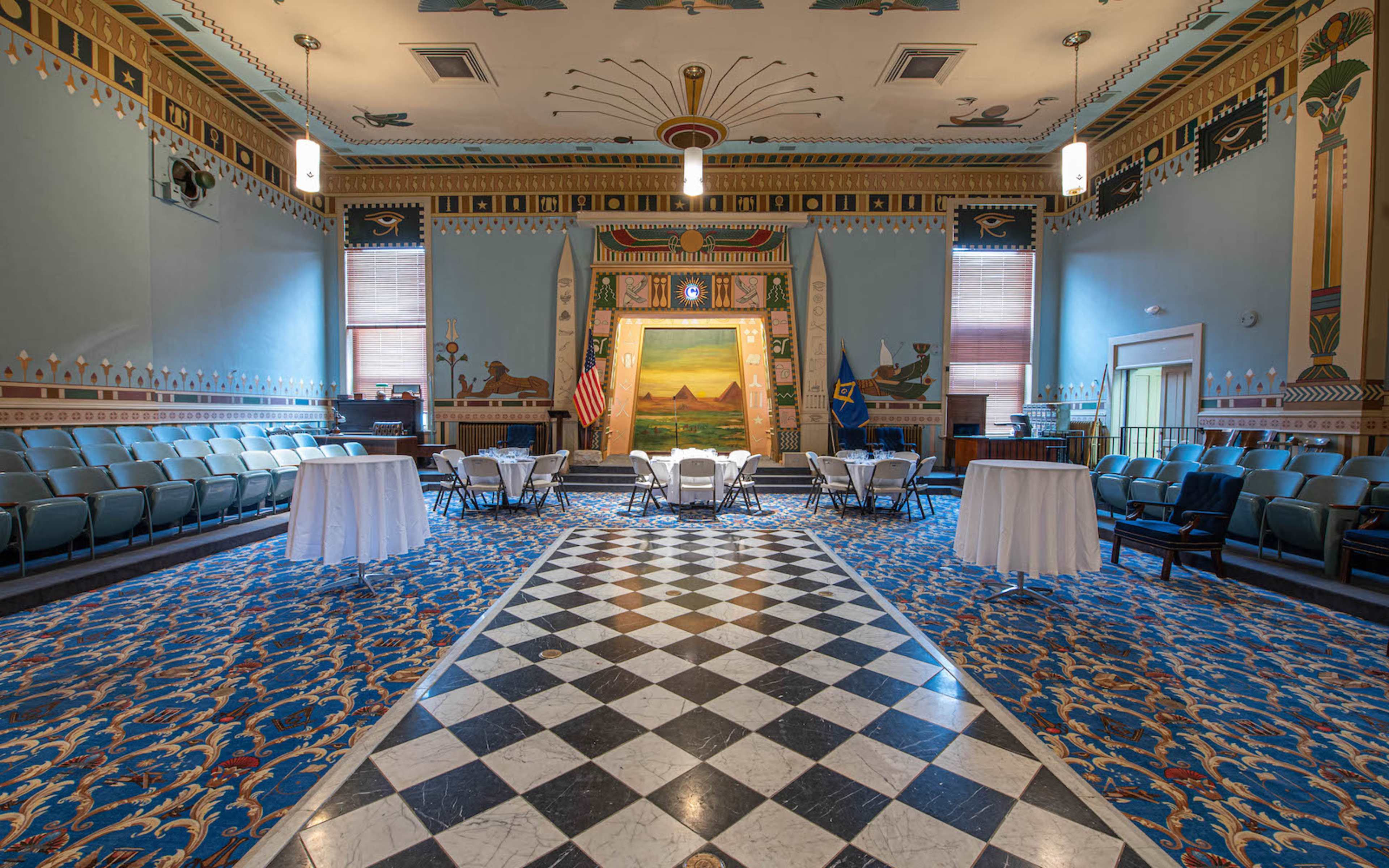 A large room with a checkered floor, featuring seating arranged in a circular pattern and tables set up in the center, adorned with decorative murals and an American flag displayed to the side.