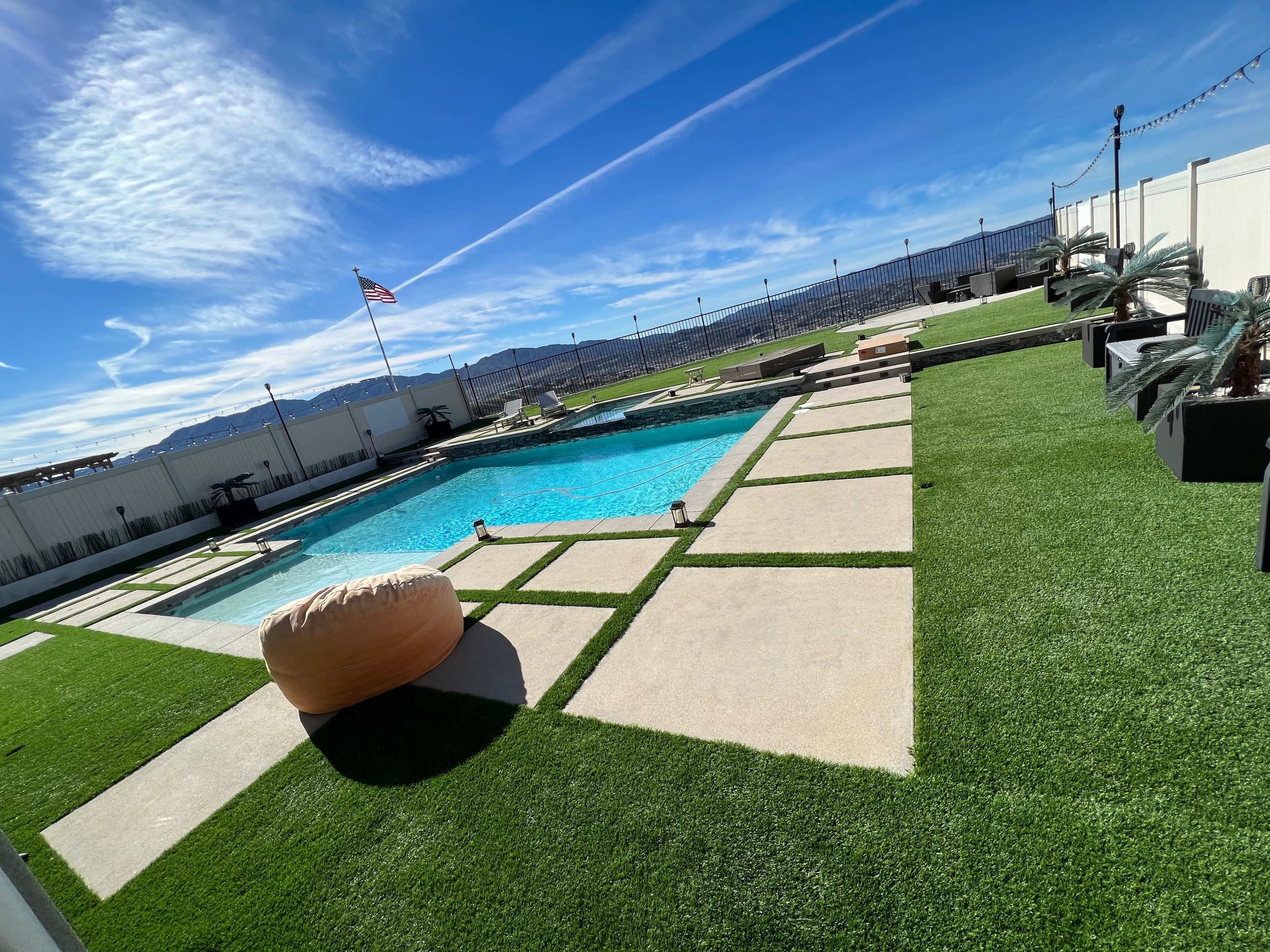 The image shows a pool area surrounded by green artificial grass, with a view of mountains in the background and an American flag flying.