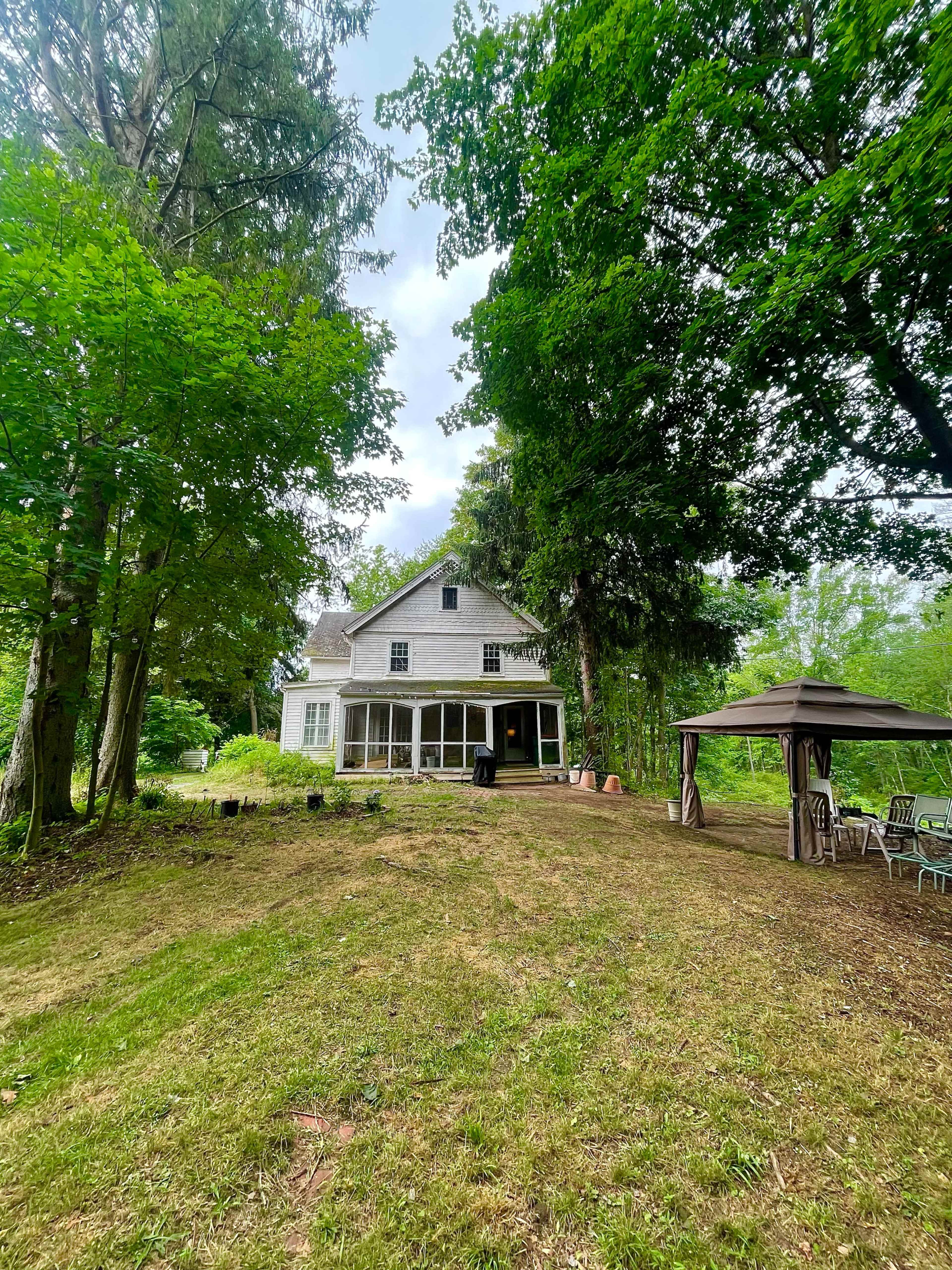 A two-story, weathered house is situated on a grassy slope, surrounded by tall trees and a gazebo nearby.