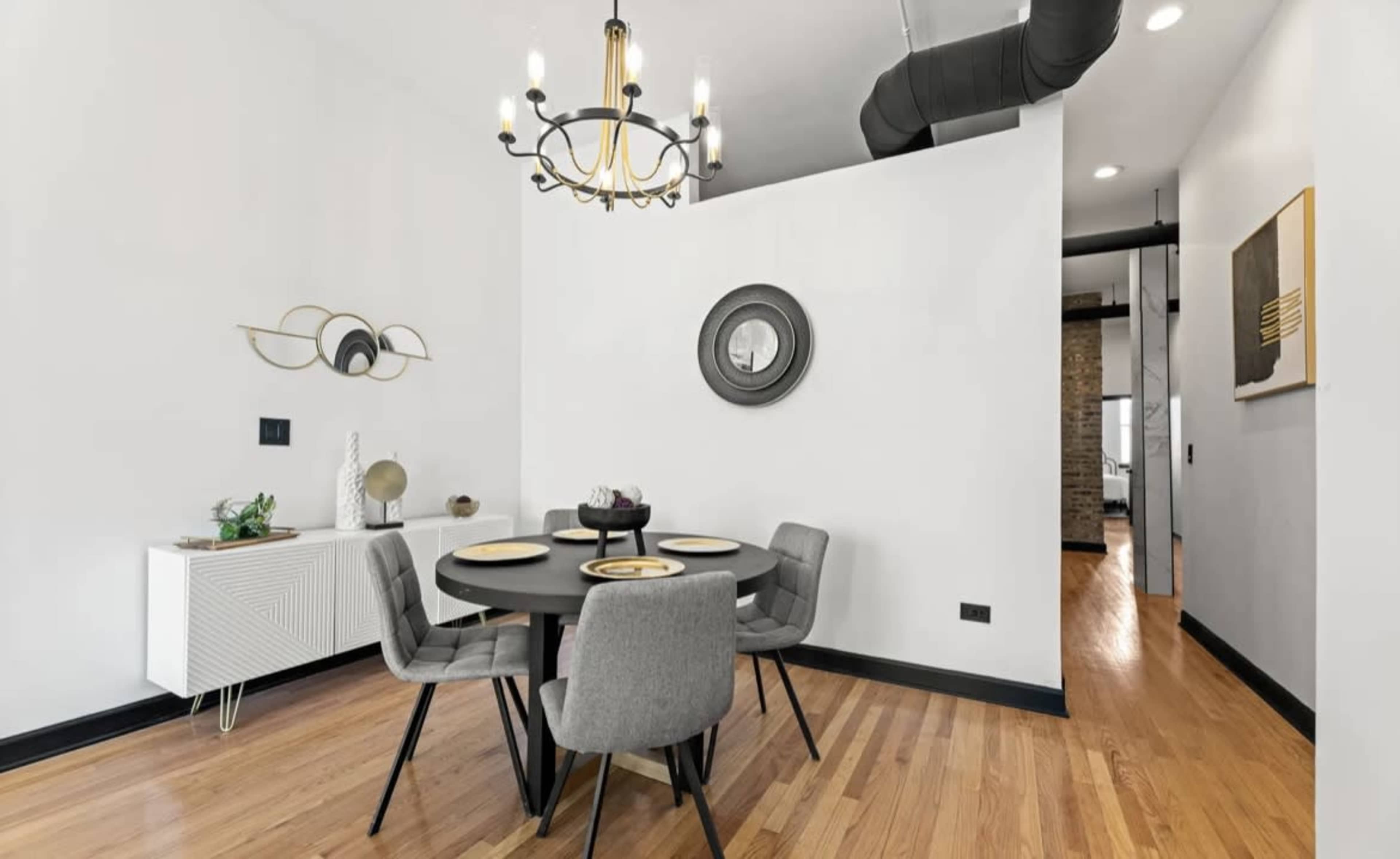 A modern dining area features a round black table set for four, surrounded by gray upholstered chairs, with a chandelier above and a sideboard against the wall.