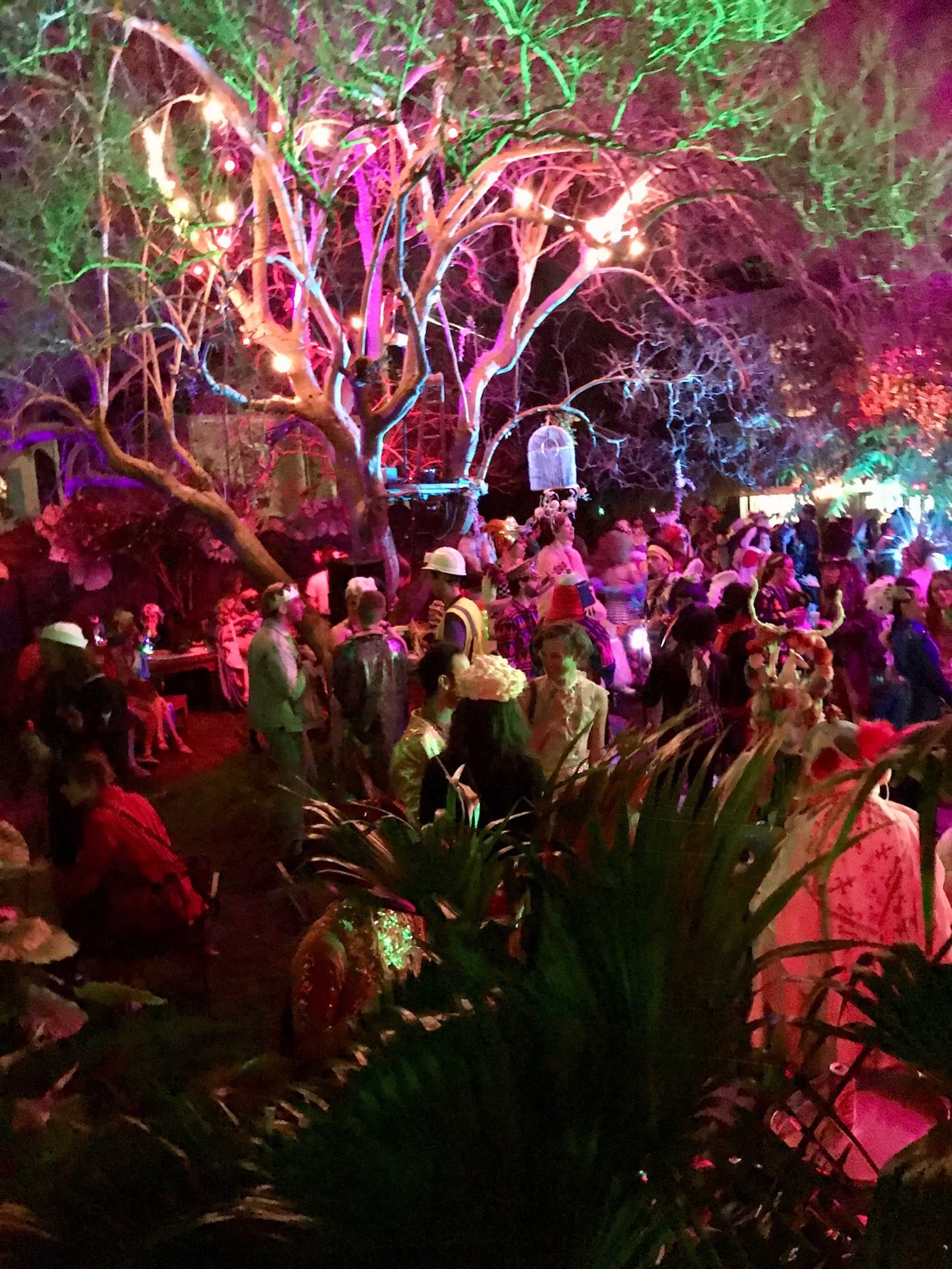 A lively gathering takes place at night under a colorful illuminated tree, with guests wearing various costumes mingling in a festive atmosphere.