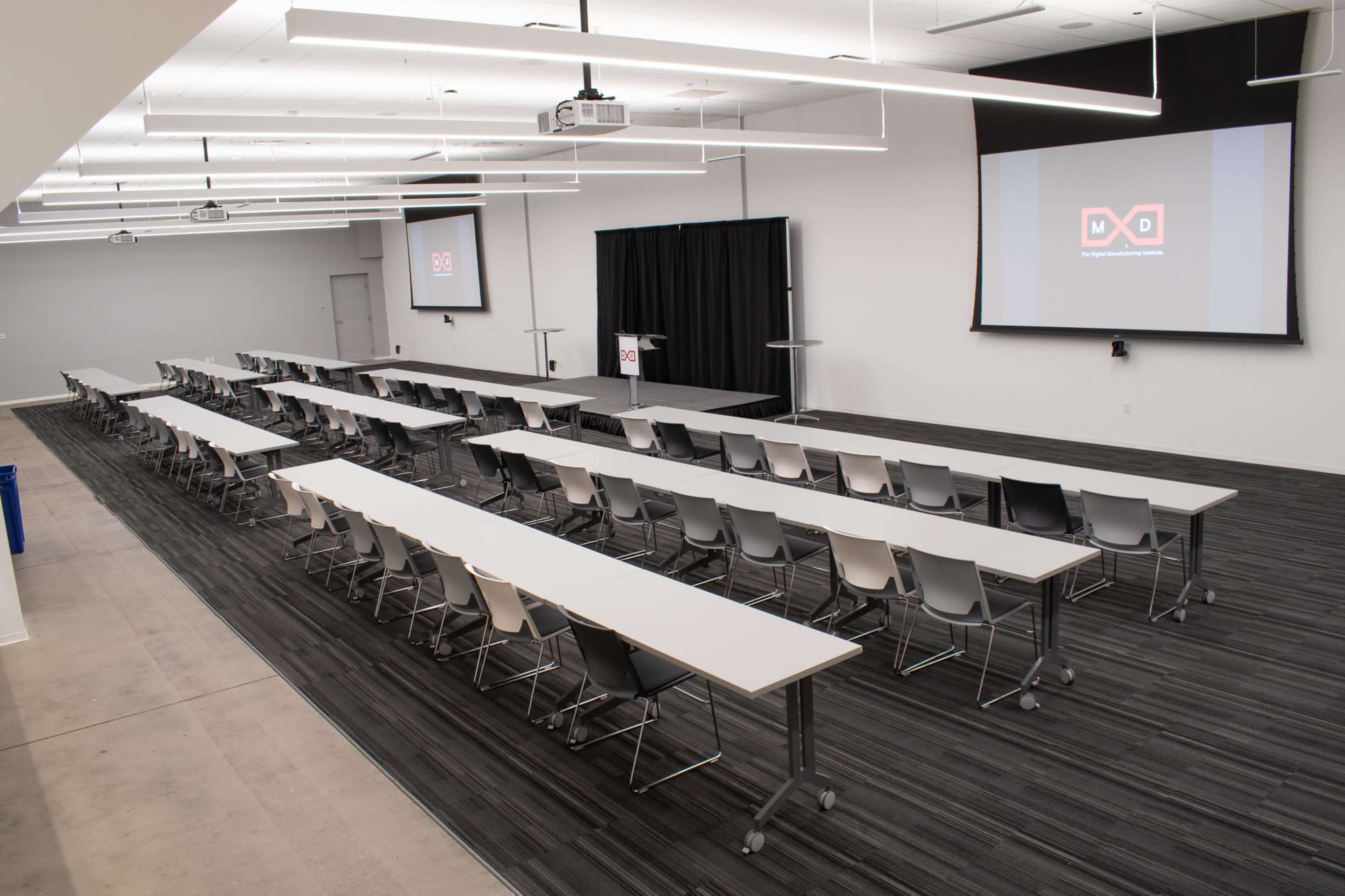 The image shows a spacious conference room arranged with multiple rows of long tables and chairs, facing a projector screen and a podium.