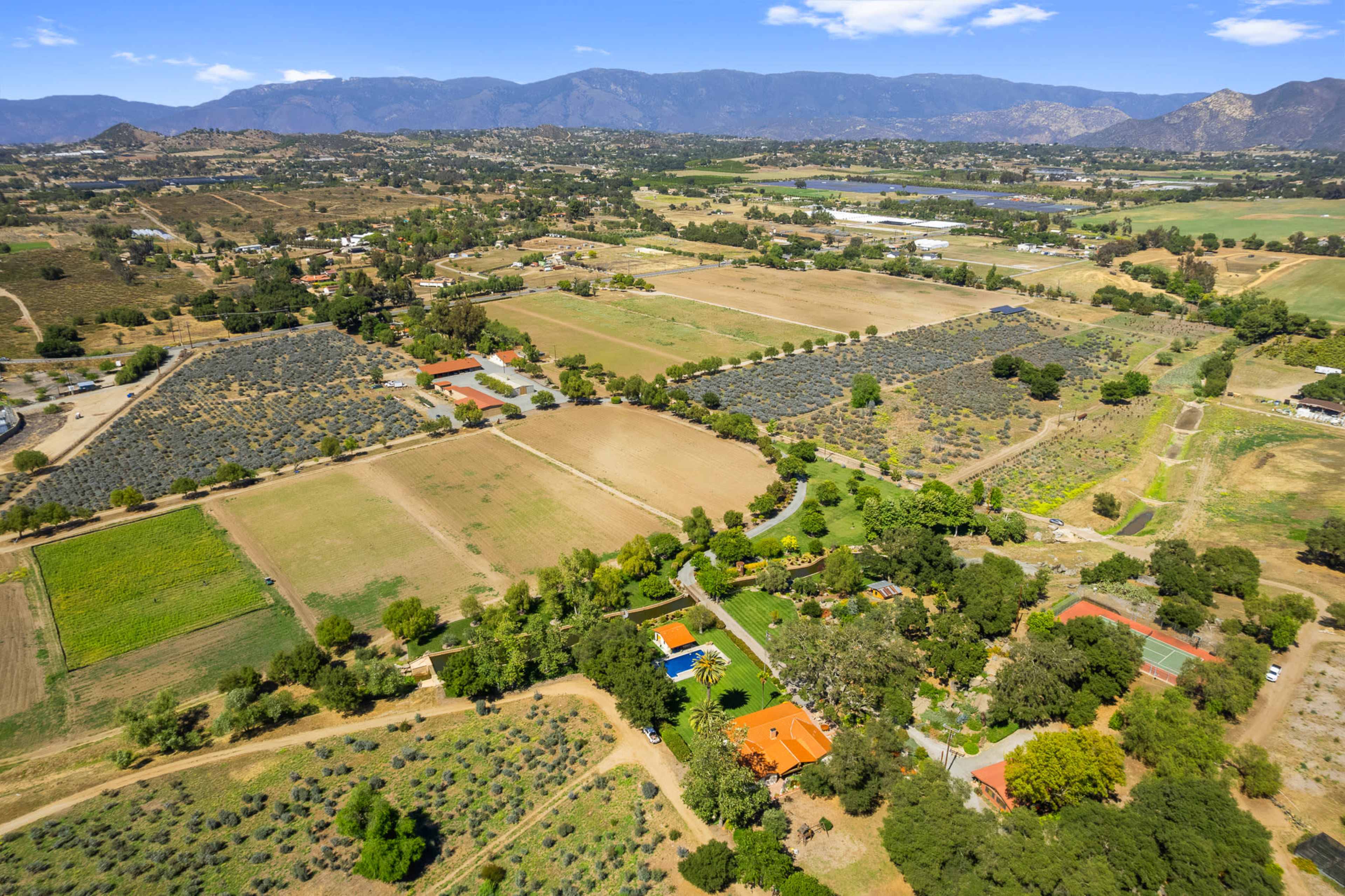 The image shows a vast agricultural landscape featuring fields, orchards, and farm buildings, surrounded by mountains in the background.