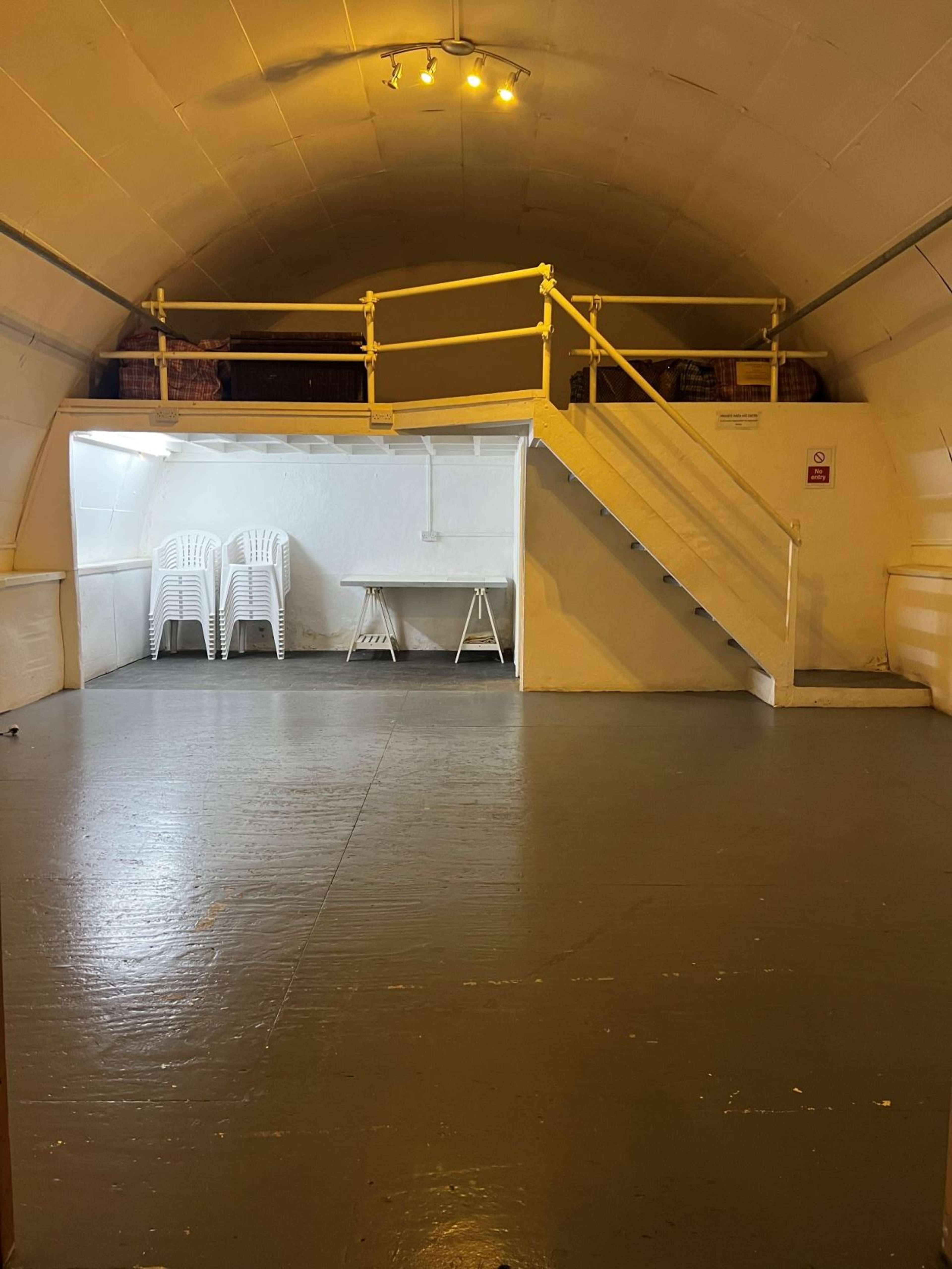 A spacious, well-lit bunker interior featuring a staircase leading to a raised platform with seating, and a table set up on the lower level.