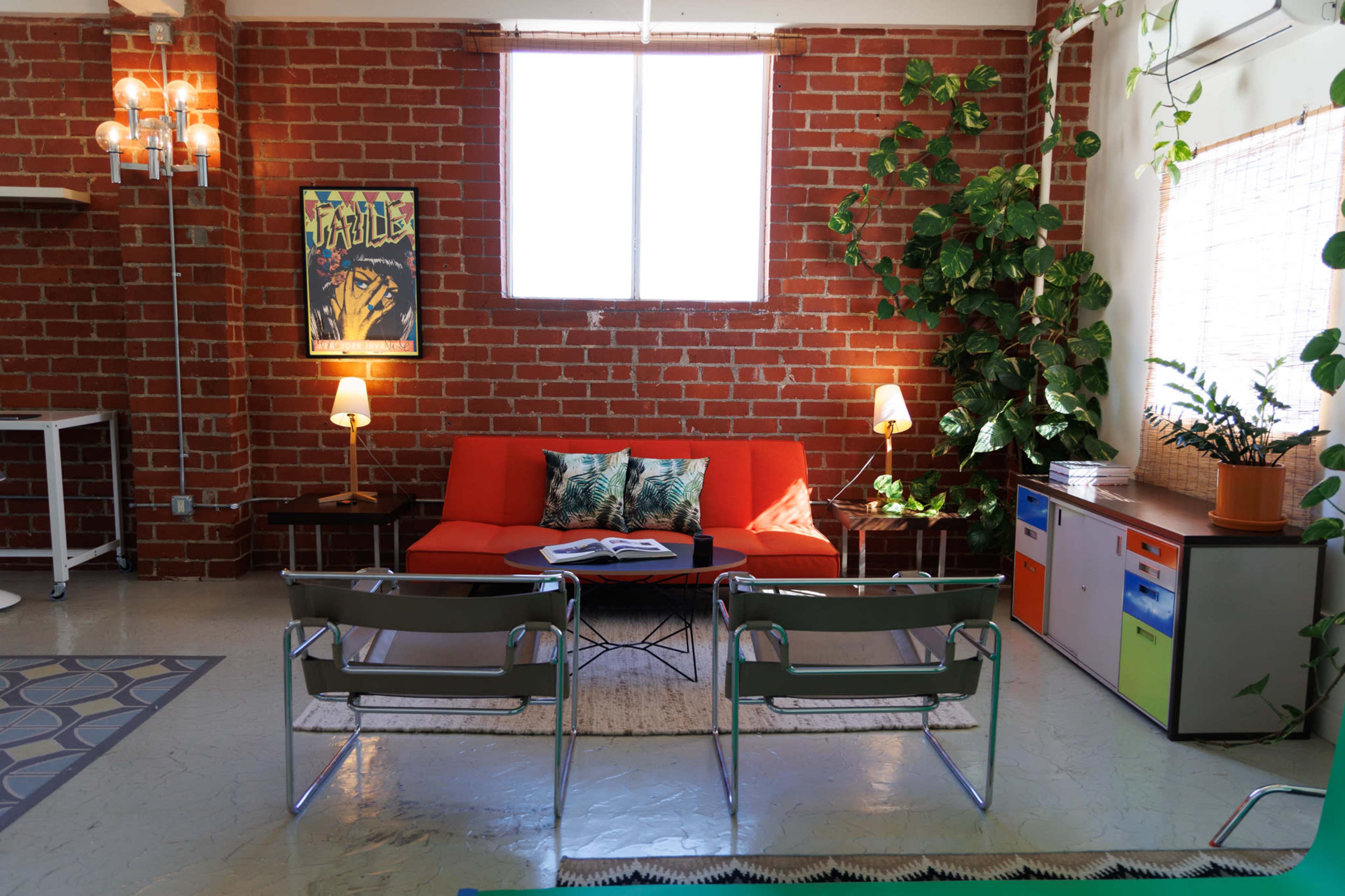 The image shows a living space with a red-brick wall, an orange couch, two chairs, a coffee table, and plants, illuminated by natural light from a window.