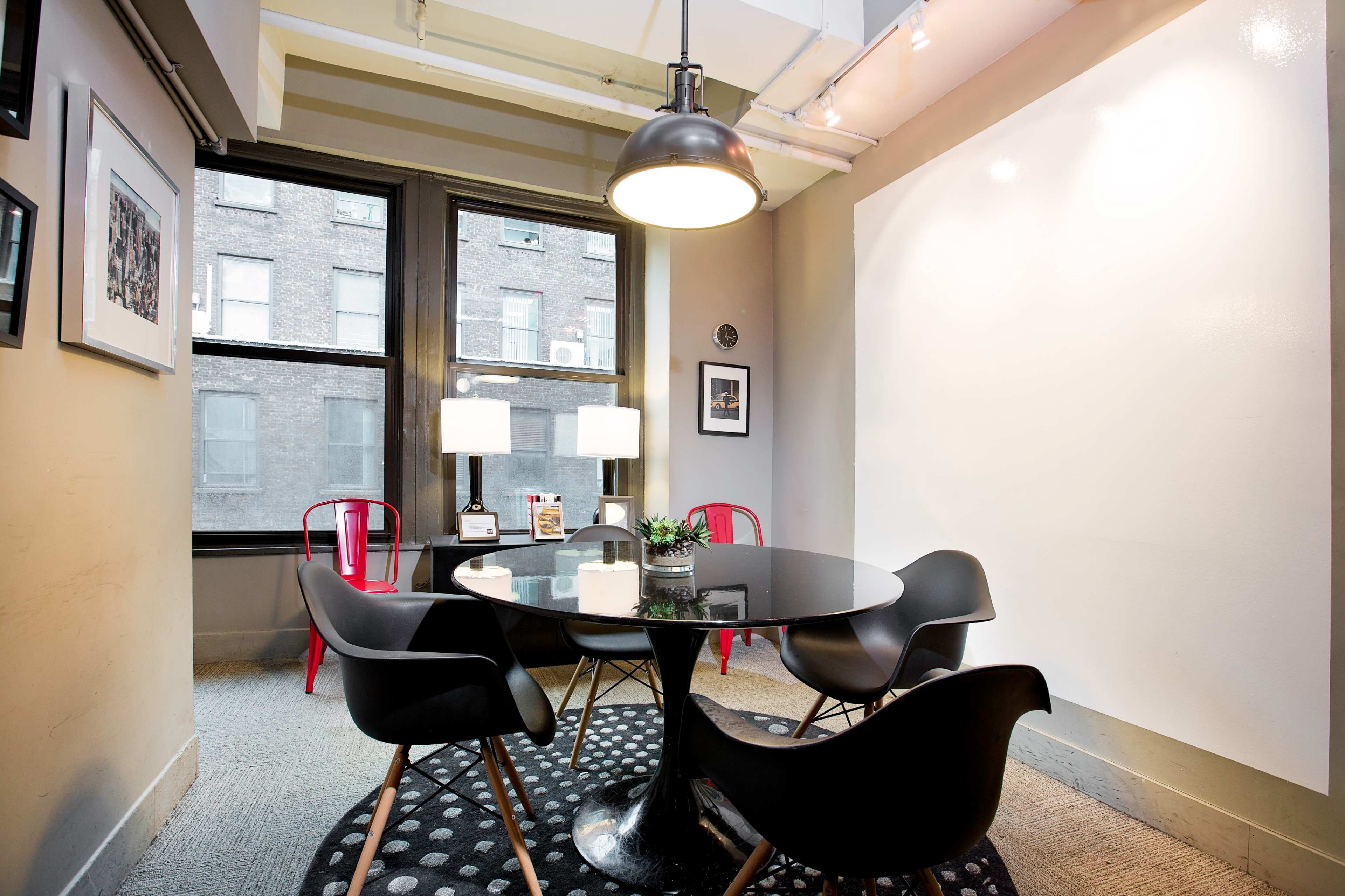 A modern small meeting room features a round black table surrounded by four chairs, with two lamps on a side table and framed pictures on the walls.