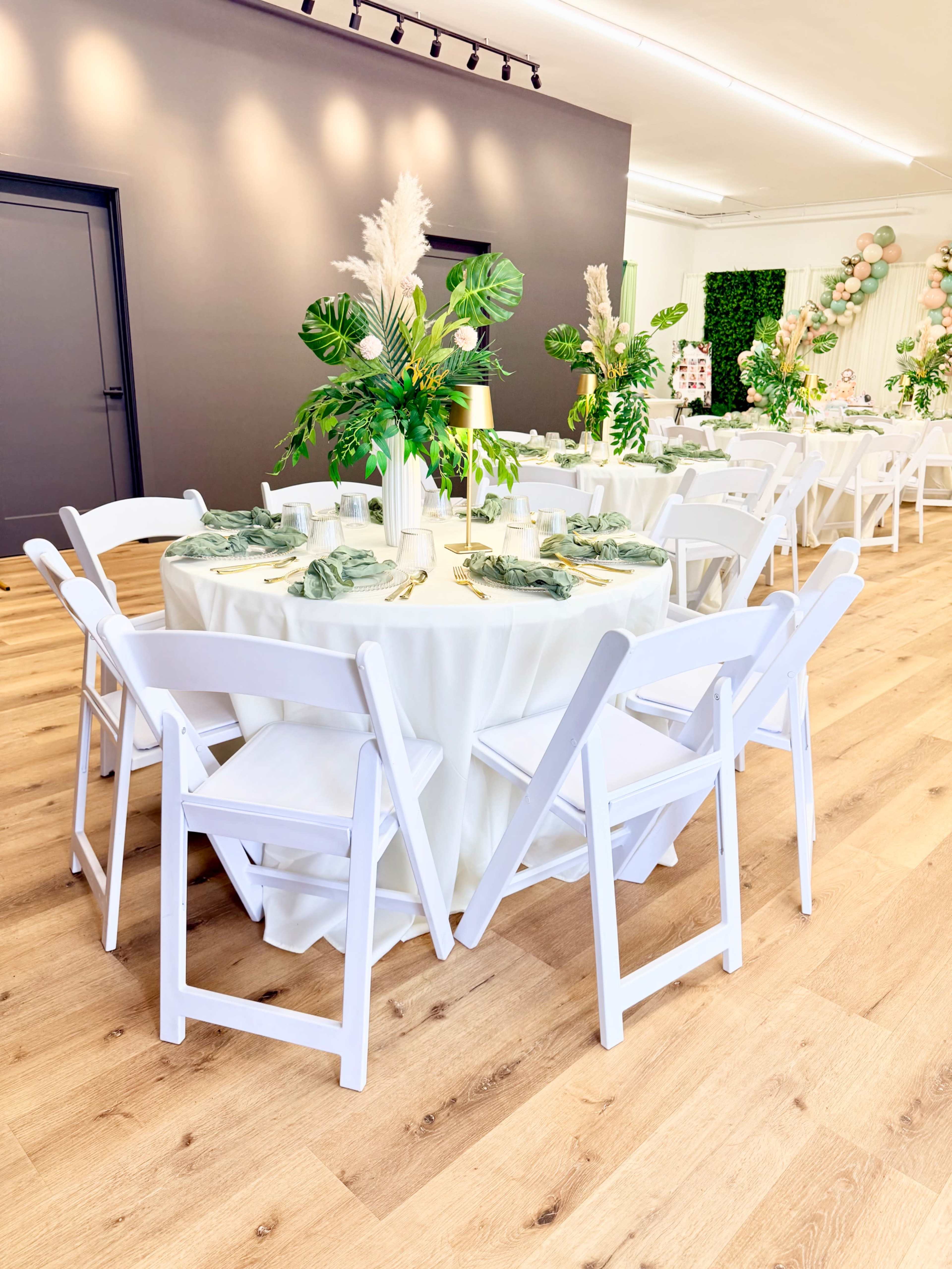 A banquet hall is set up with round tables covered in white tablecloths, surrounded by white chairs, and adorned with green plants and decorative elements.