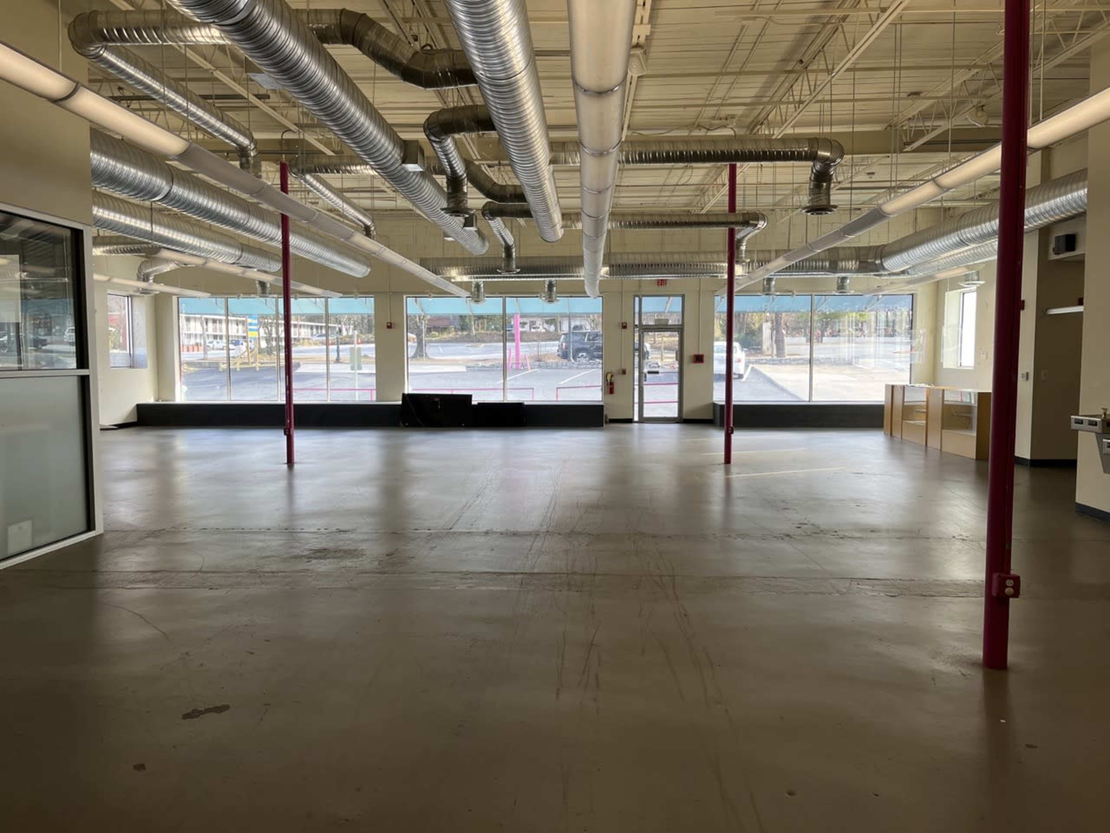 An empty commercial space with large windows, exposed ductwork, and a concrete floor.