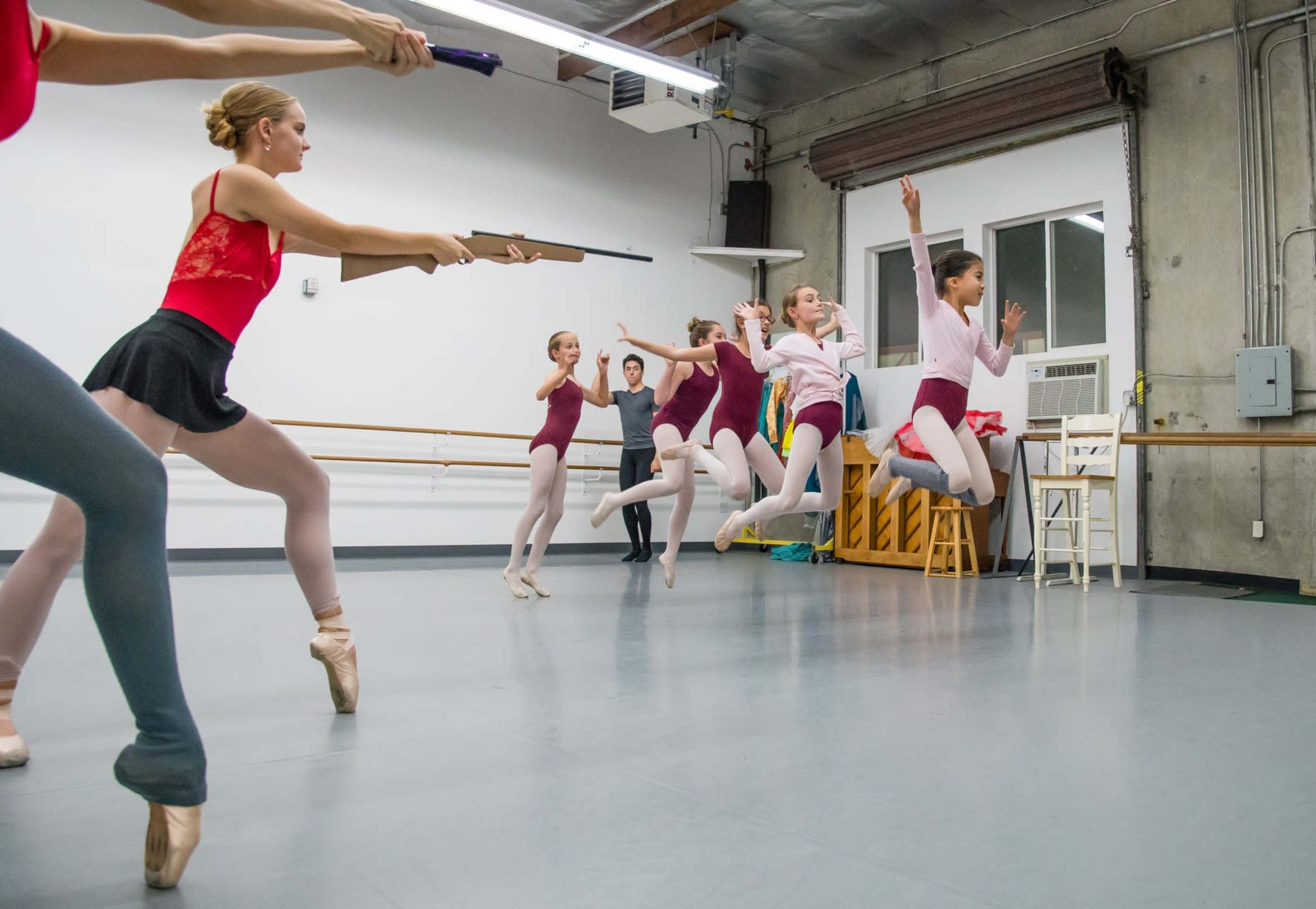 A group of ballet dancers practices in a studio, some jumping while others hold positions, with an instructor observing.