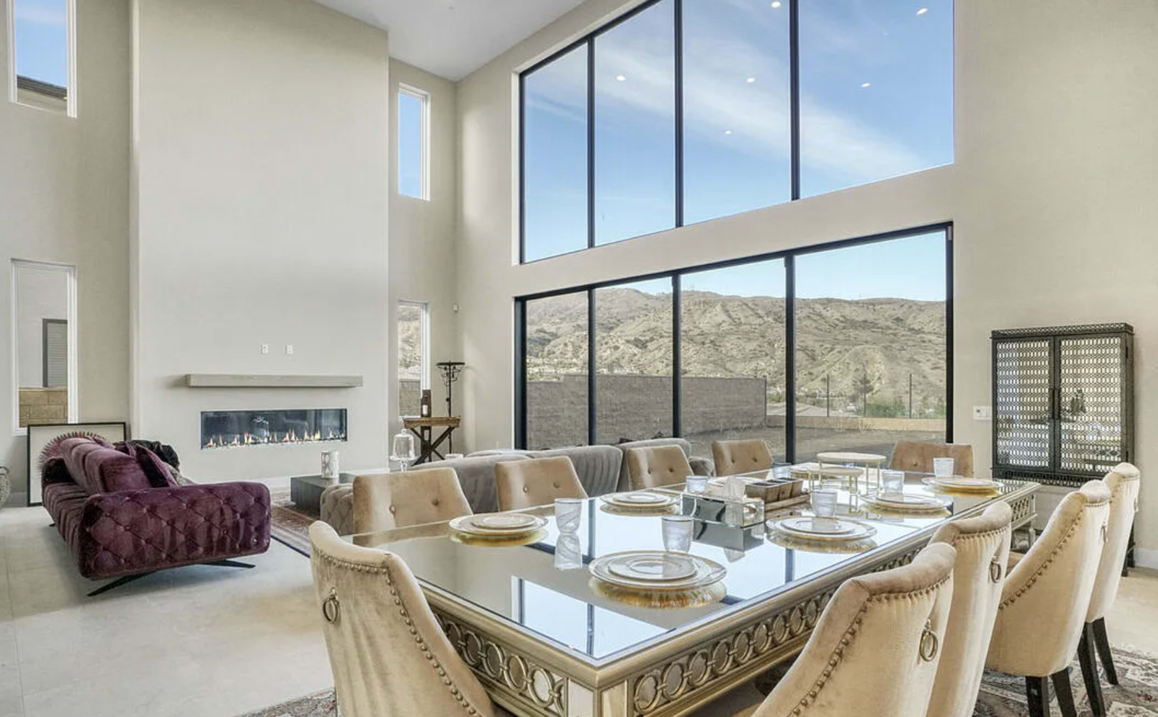 A spacious dining area features a long table set for a meal, with large windows showcasing a mountainous view and a modern fireplace along one wall.