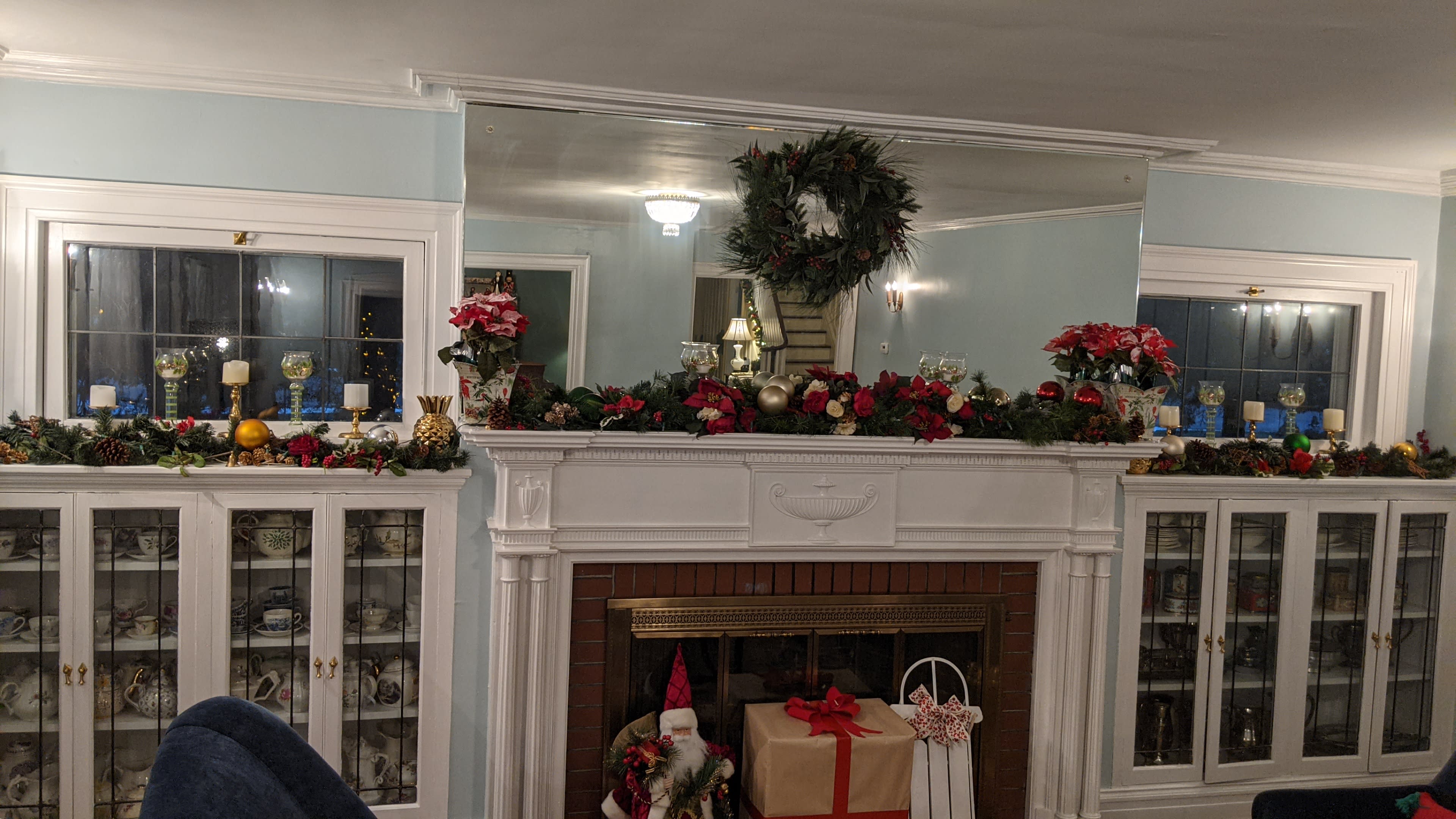 The image shows a decorated fireplace mantel adorned with garlands, red flowers, and holiday ornaments, set against a light blue wall with matching decor in the background.