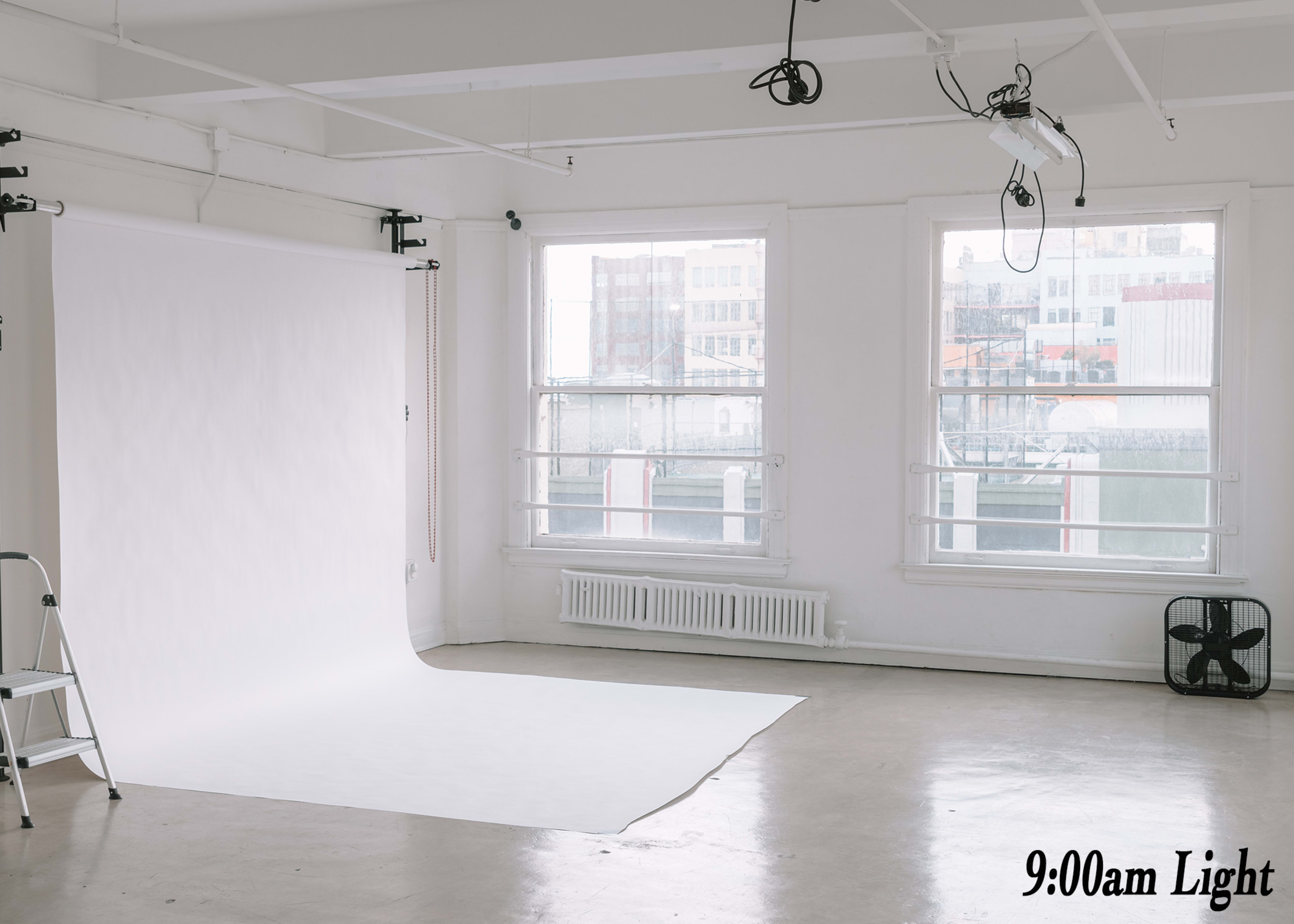 The image shows a bright photography studio with a white backdrop, a ladder, and large windows allowing natural light to illuminate the space.