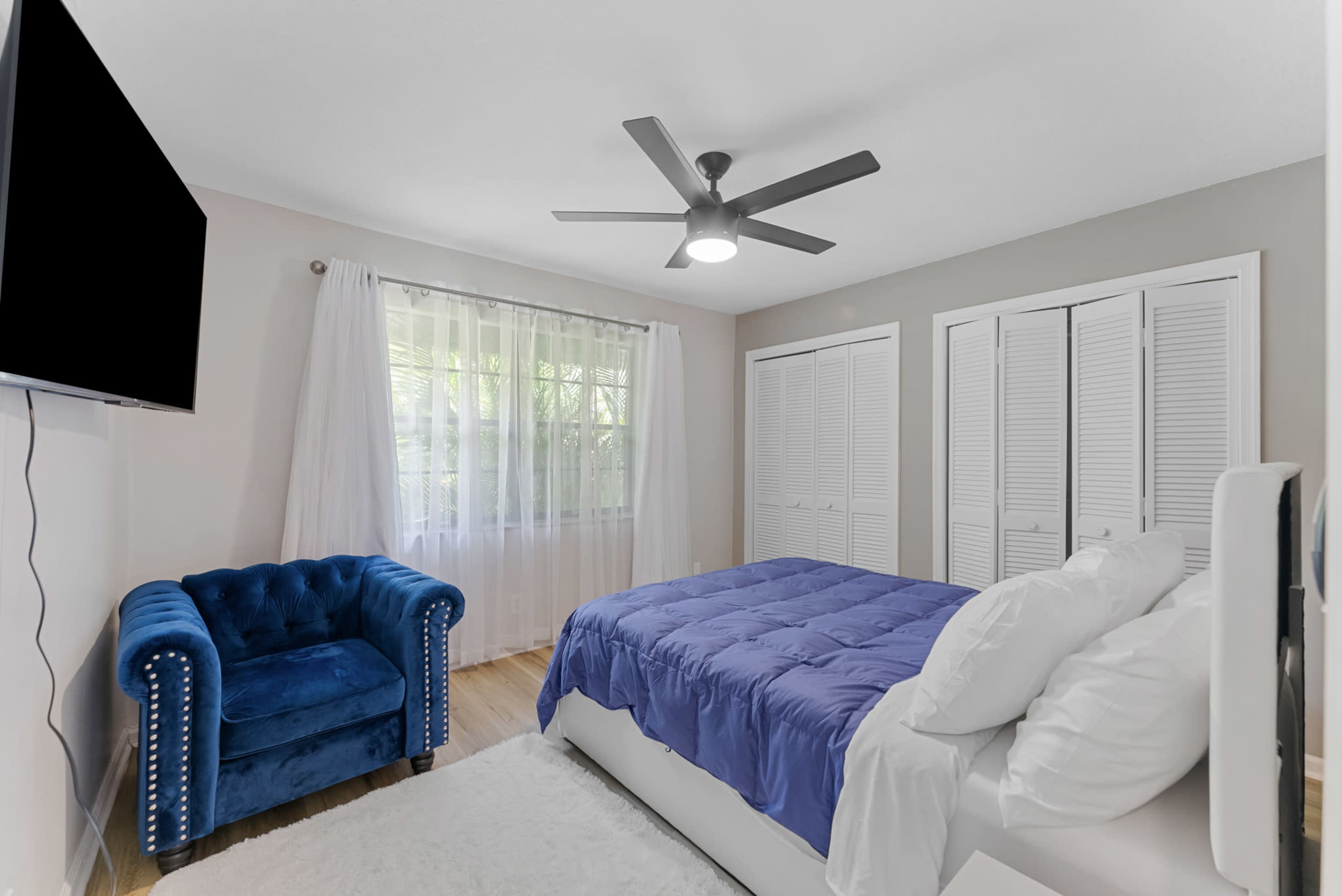 A modern bedroom features a bed with a blue comforter, a blue armchair, a large wall-mounted TV, and white closet doors.