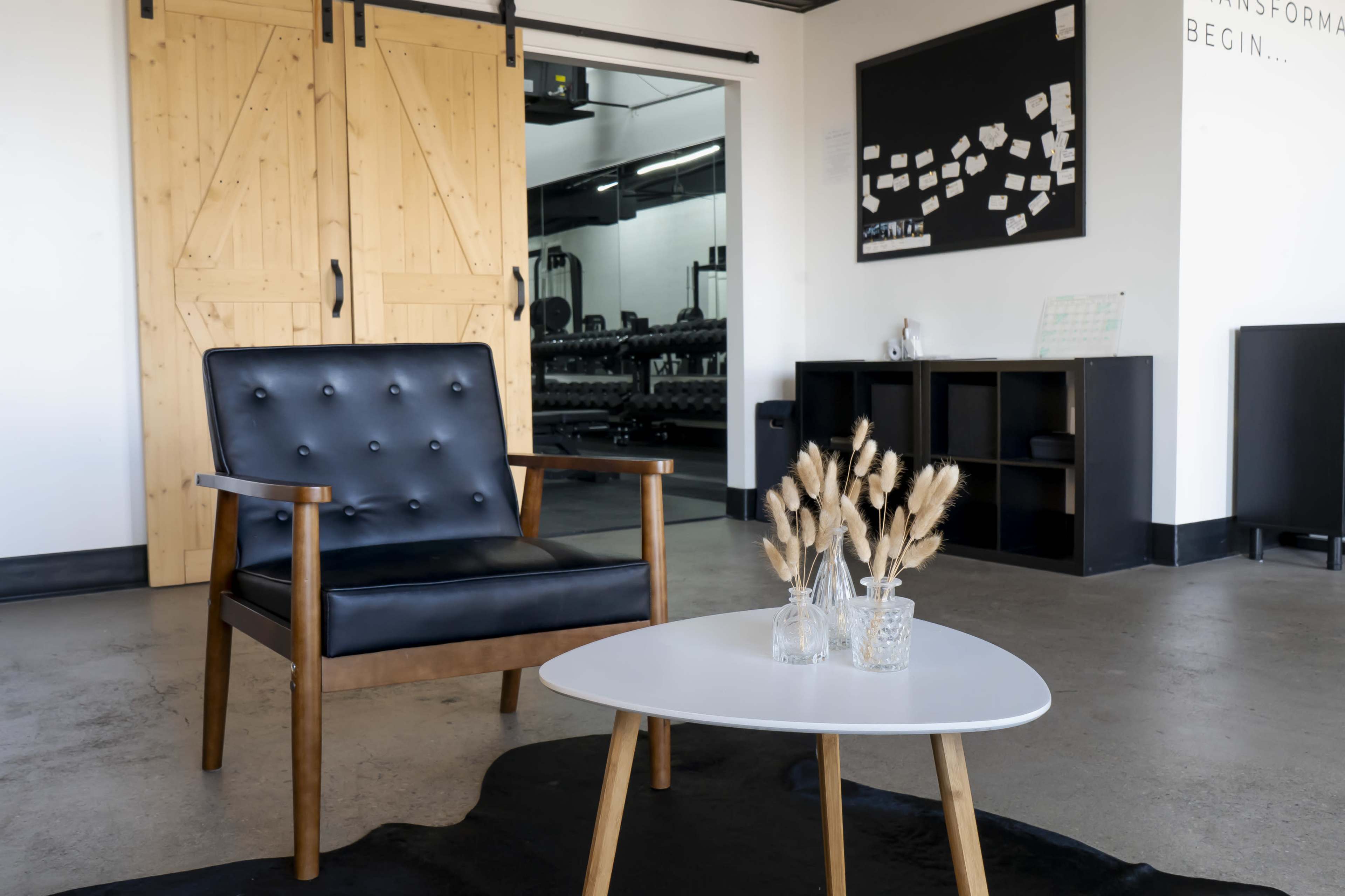 The image shows a modern interior with a black armchair, a white round table with a vase of dried flowers, and large wooden doors leading to a gym area.