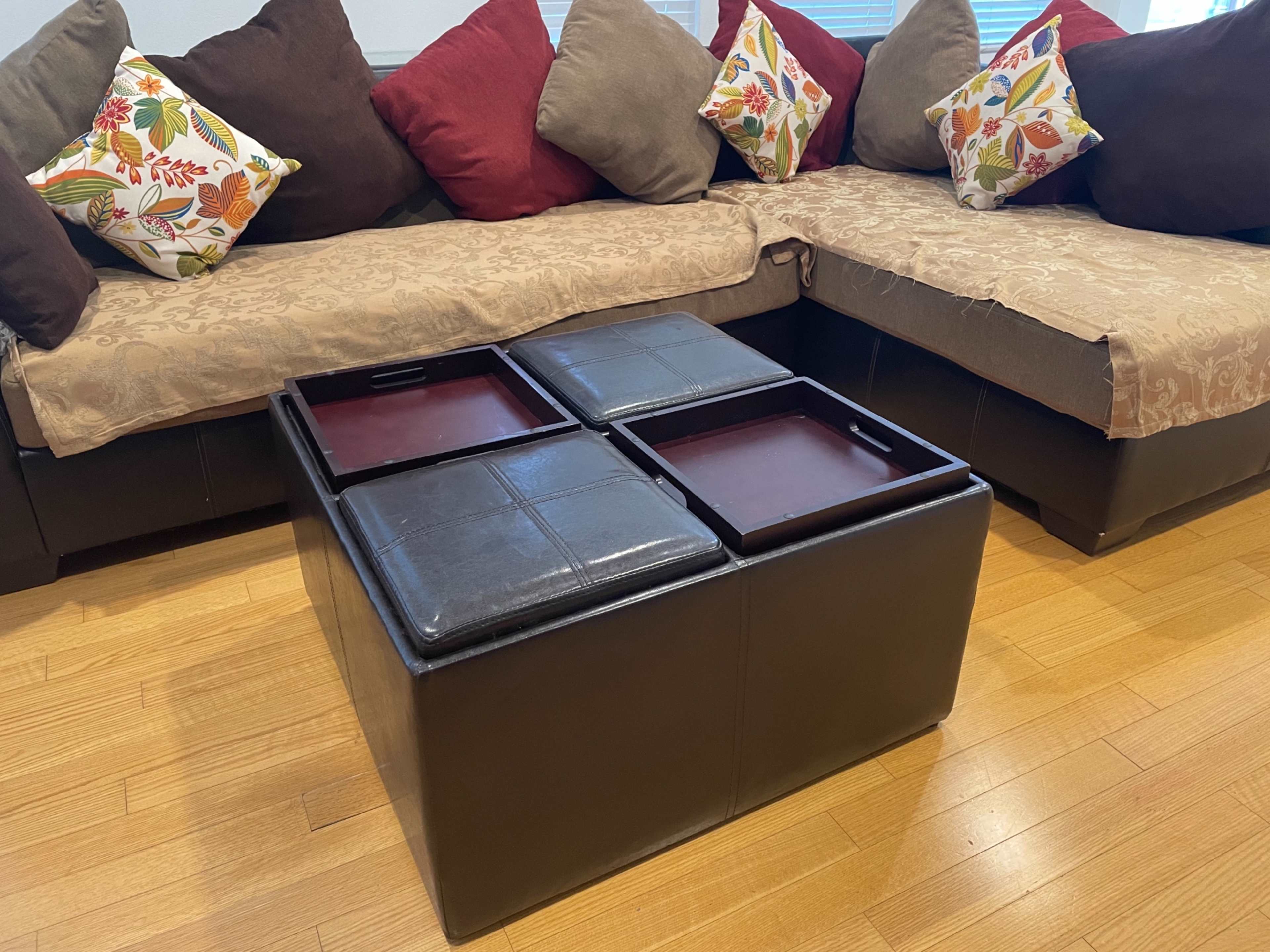 A modern living room setup featuring a brown leather ottoman with four removable top sections, surrounded by a sectional sofa with decorative pillows.