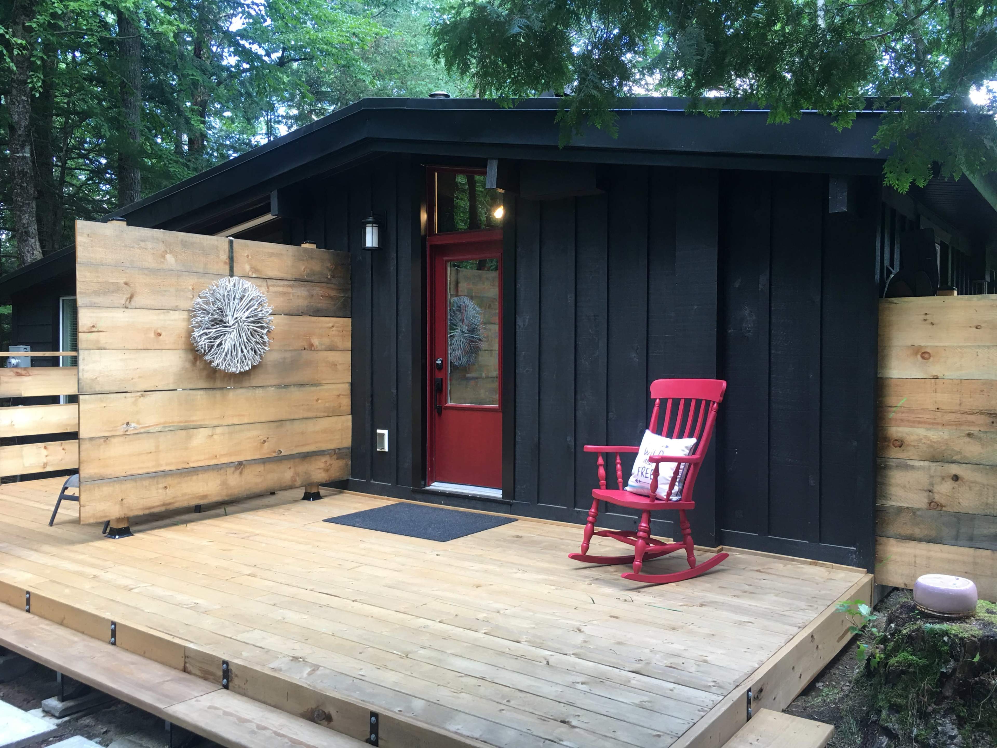 A modern wooden house with a red door and a rocking chair on a wooden deck surrounded by trees.