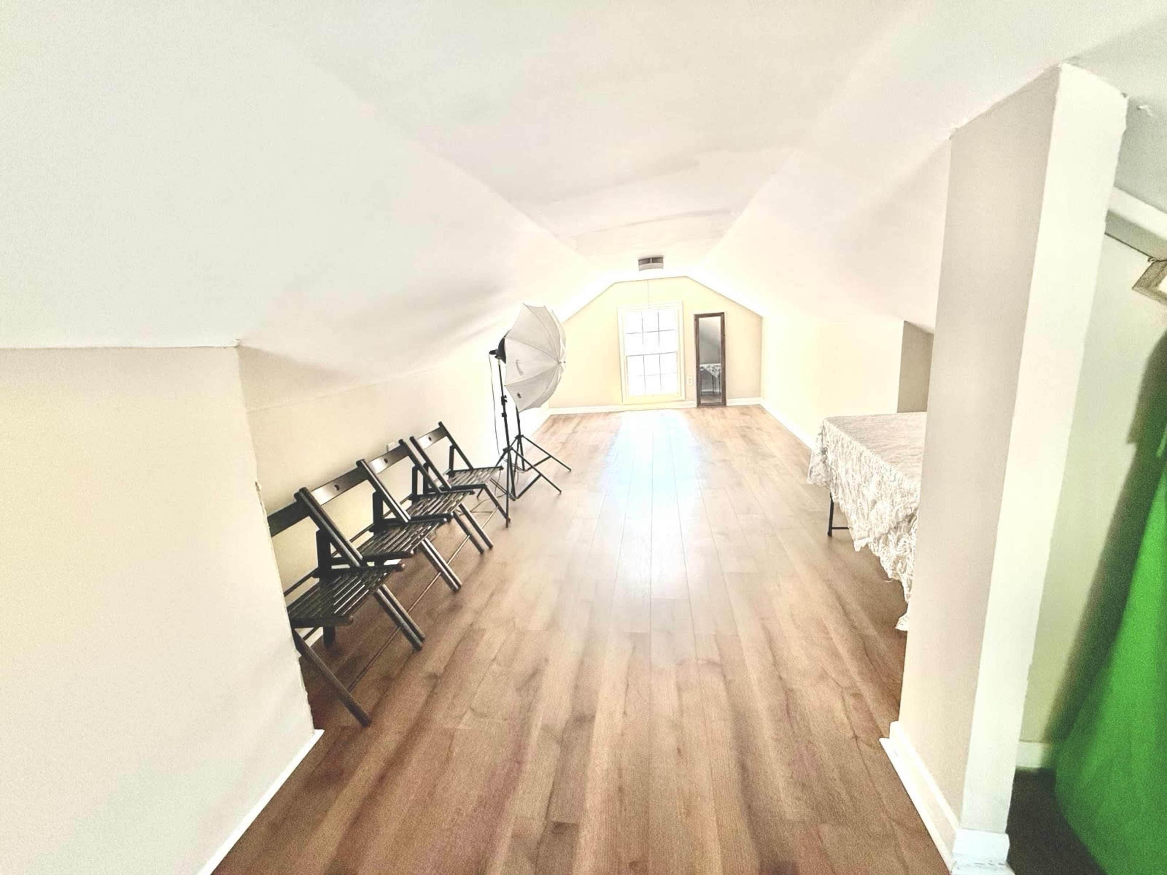 The image displays an attic space featuring a row of black chairs on the left, a large light modifier in the center, and a table covered with a light-colored cloth on the right.