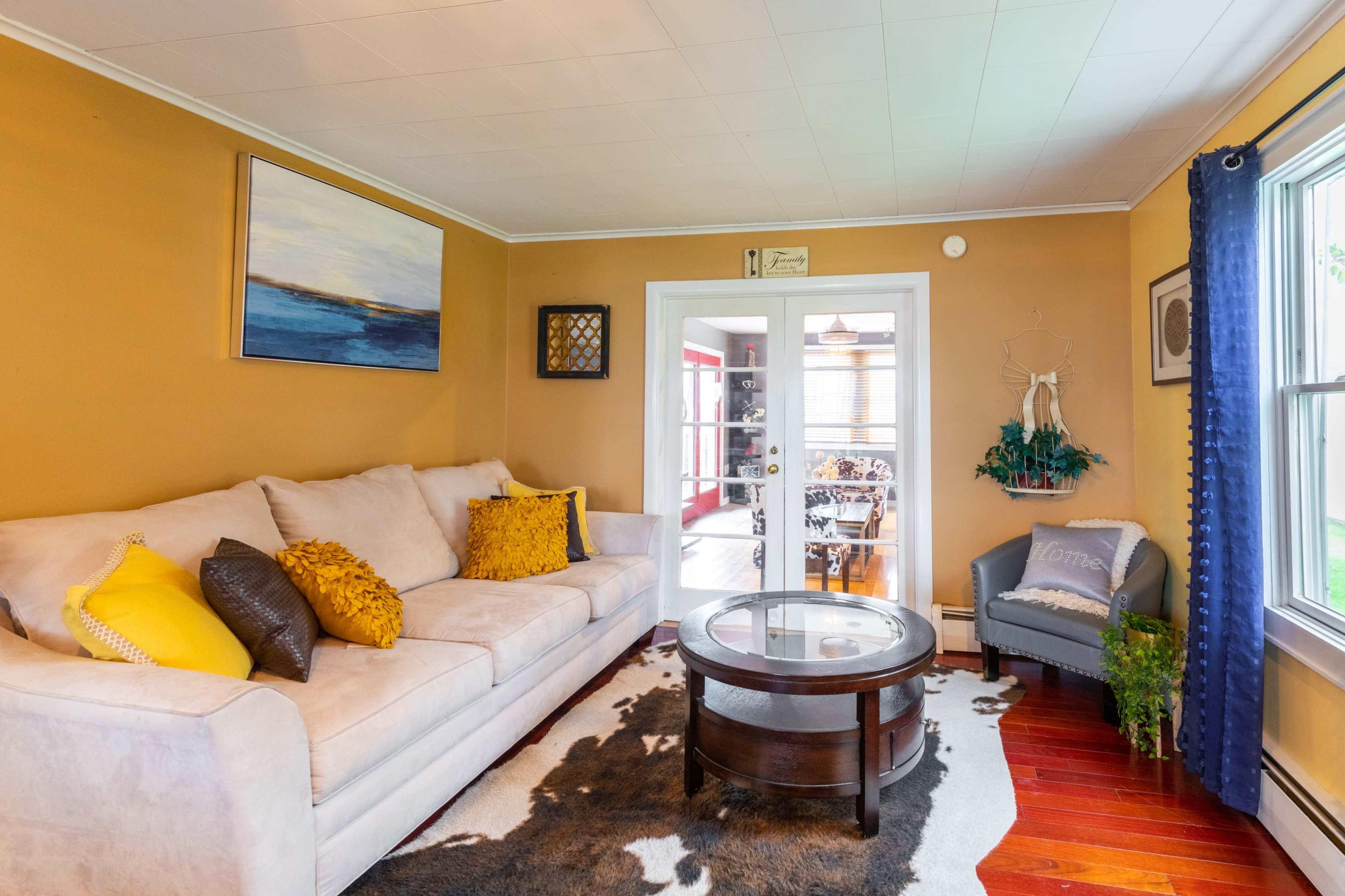 The image shows a cozy living room with a beige sofa, a round coffee table, and a large window letting in natural light.