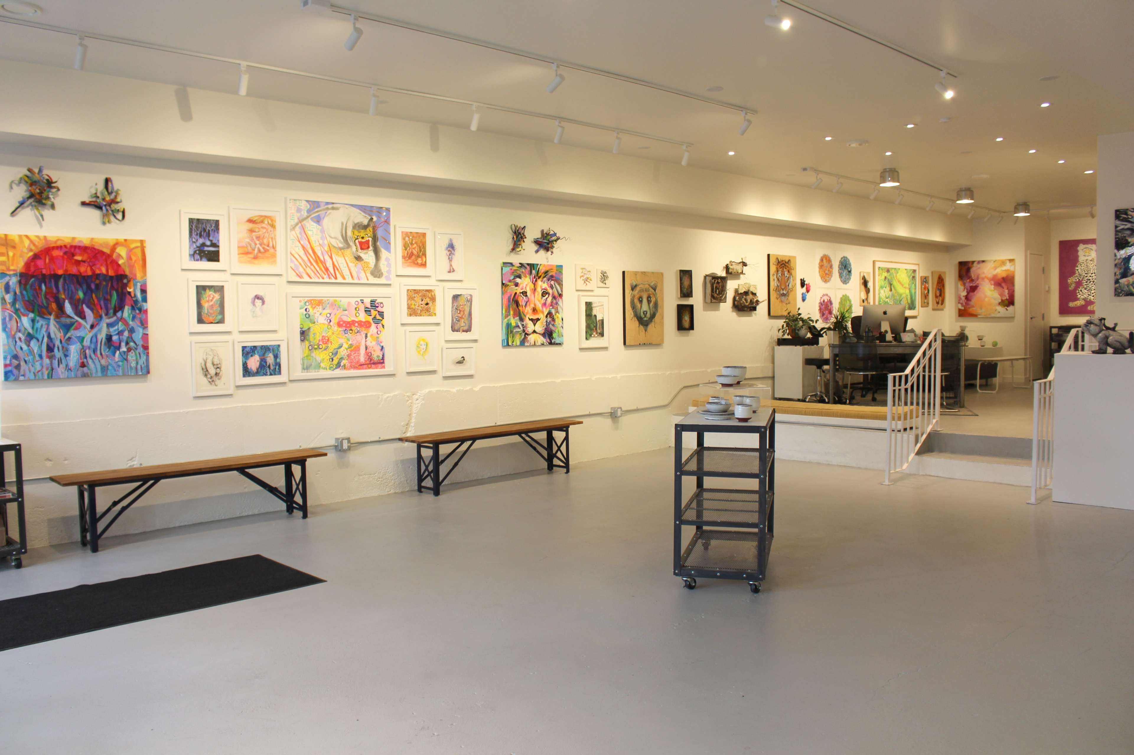An art gallery with a variety of colorful artworks displayed on the white walls and a seating area with benches along one side.