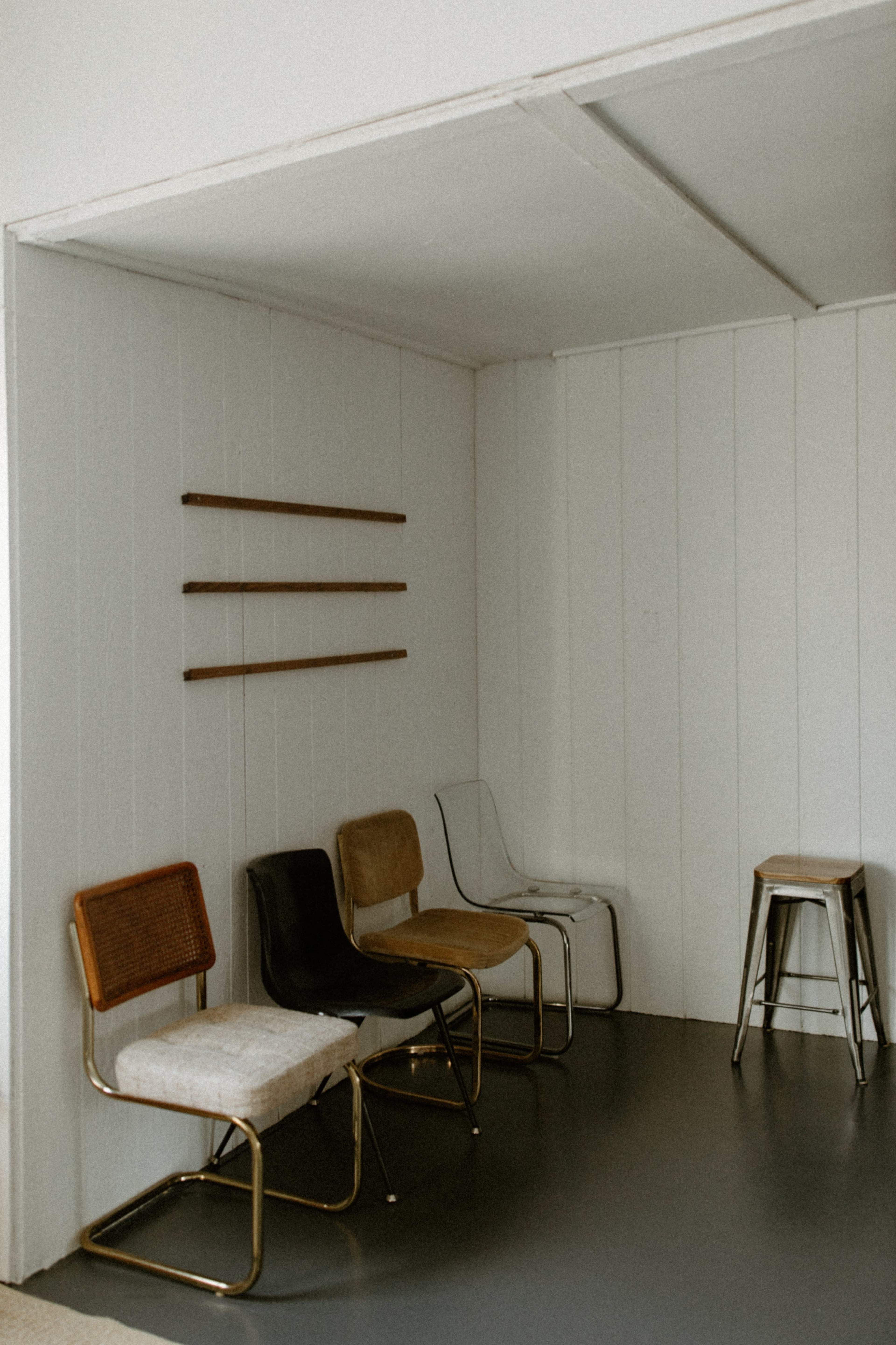 A minimalist room with a concrete floor and five chairs of varying styles arranged against a white wall.
