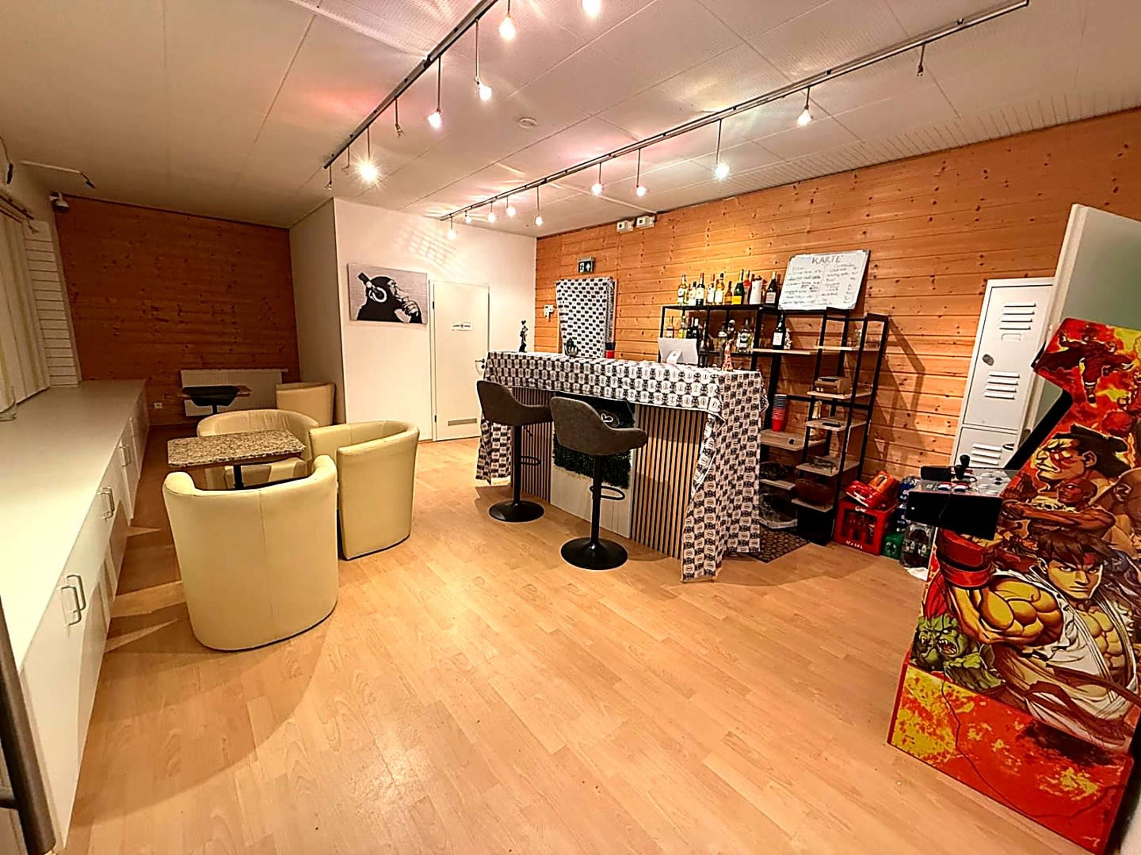 The image shows a modern interior of a bar area featuring a counter with various bottles, seating arrangements, and a video game arcade cabinet in the corner.