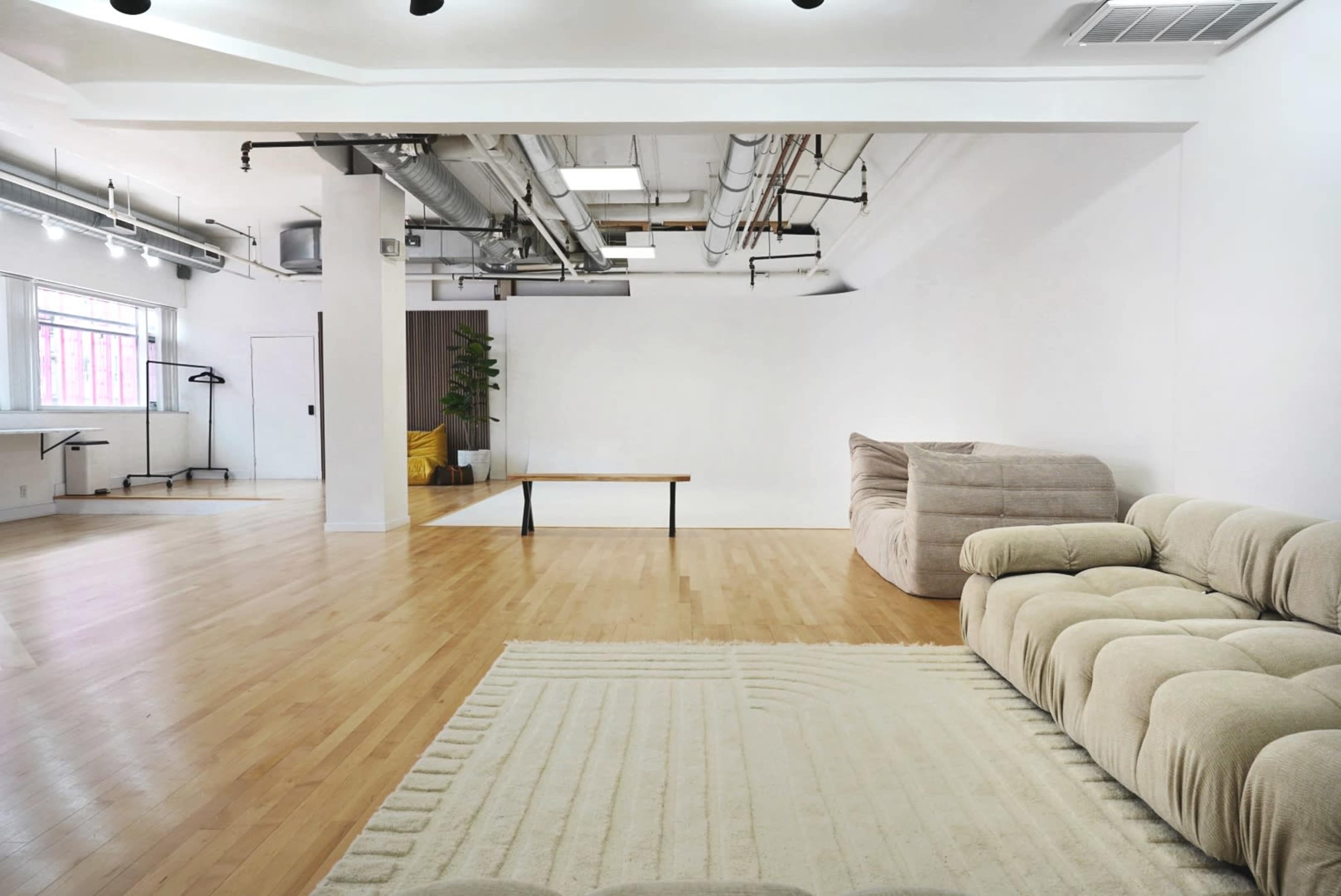 A spacious, minimalistic room with wooden flooring, a large couch, a wooden table, and large windows allowing natural light.