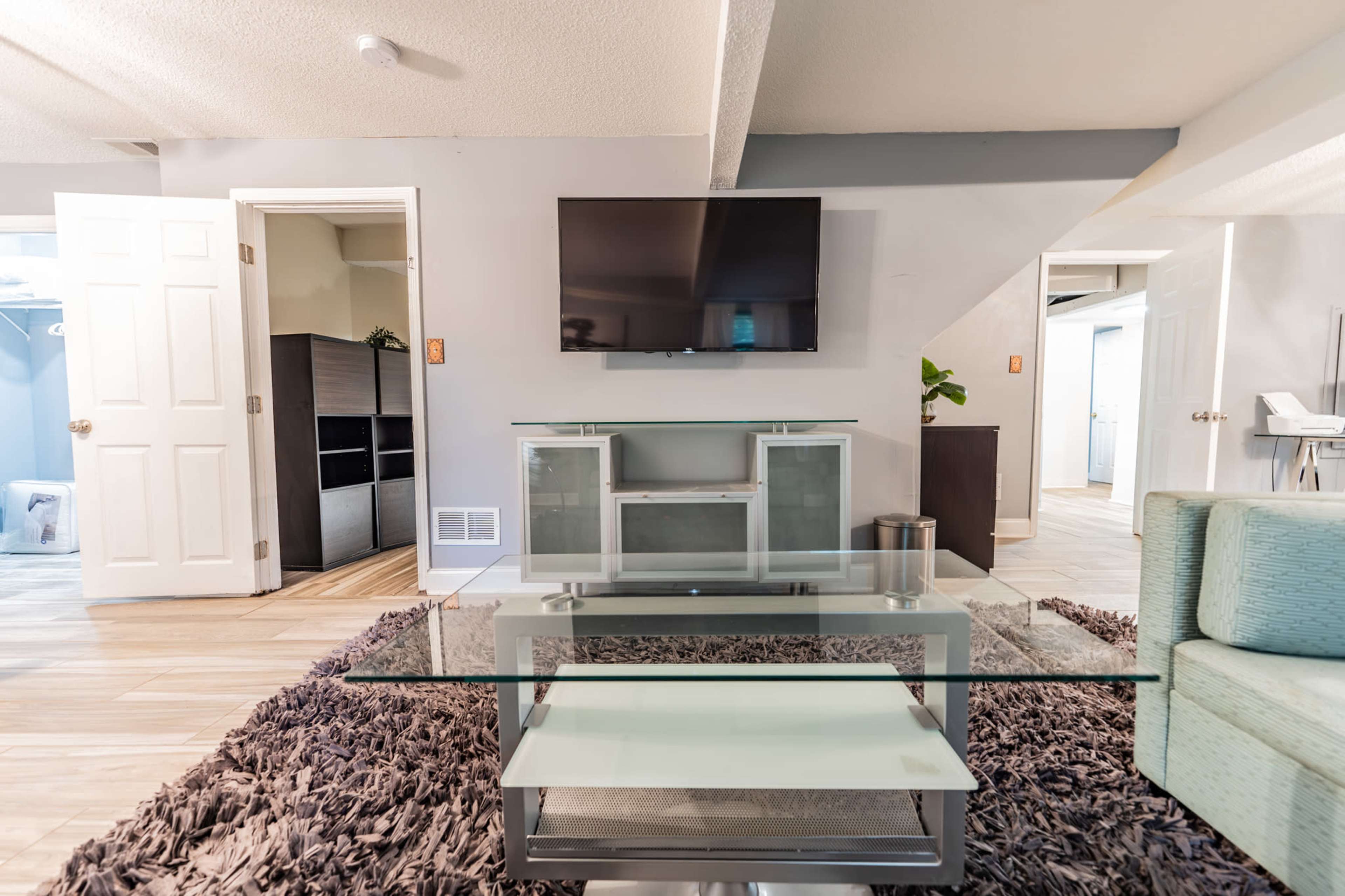The image shows a modern living room with a glass coffee table, a flat-screen TV mounted on the wall, and open doors leading to other rooms.