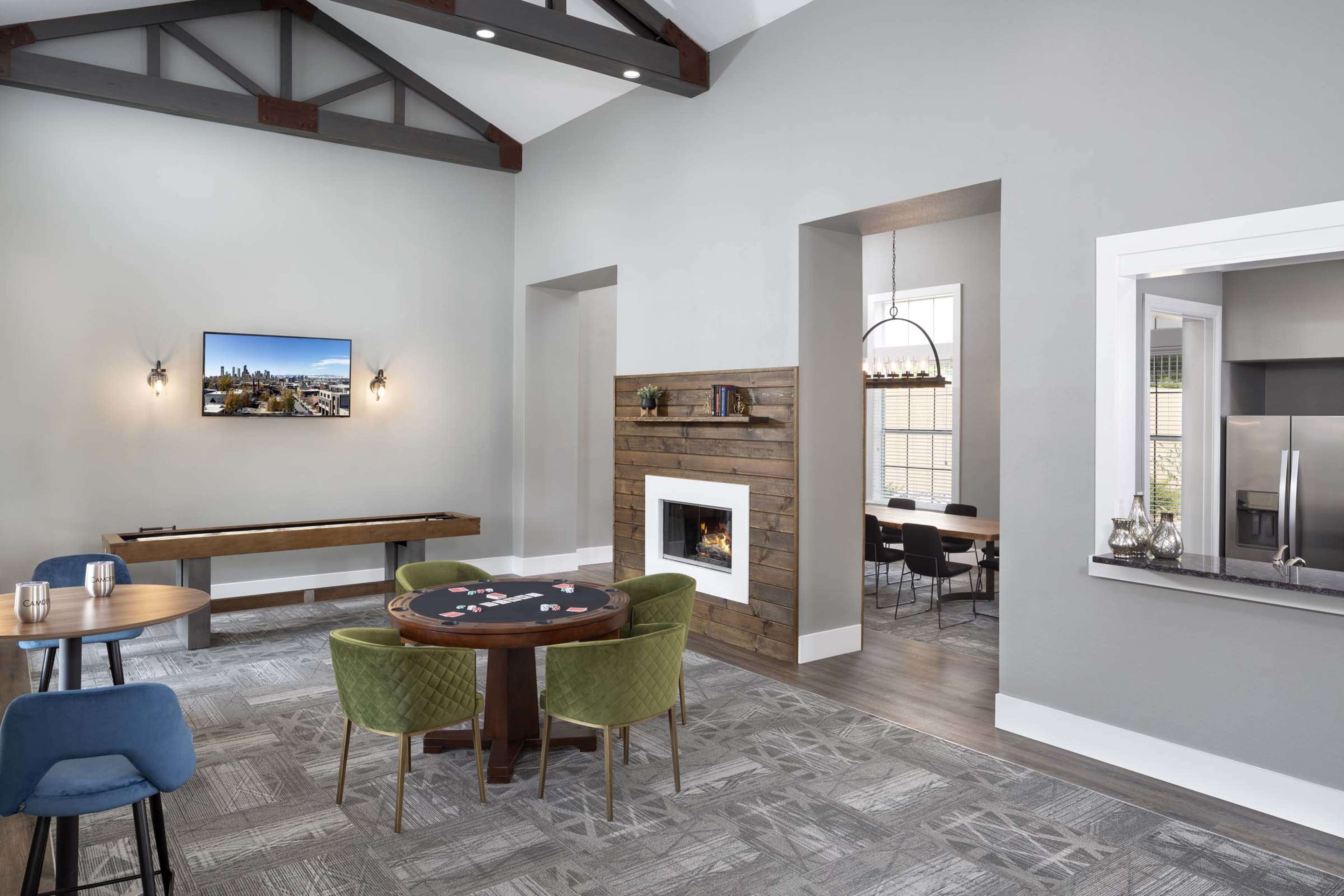 A modern communal space featuring a poker table, a dining area, and a view of a fireplace, with a large window and a television mounted on the wall.