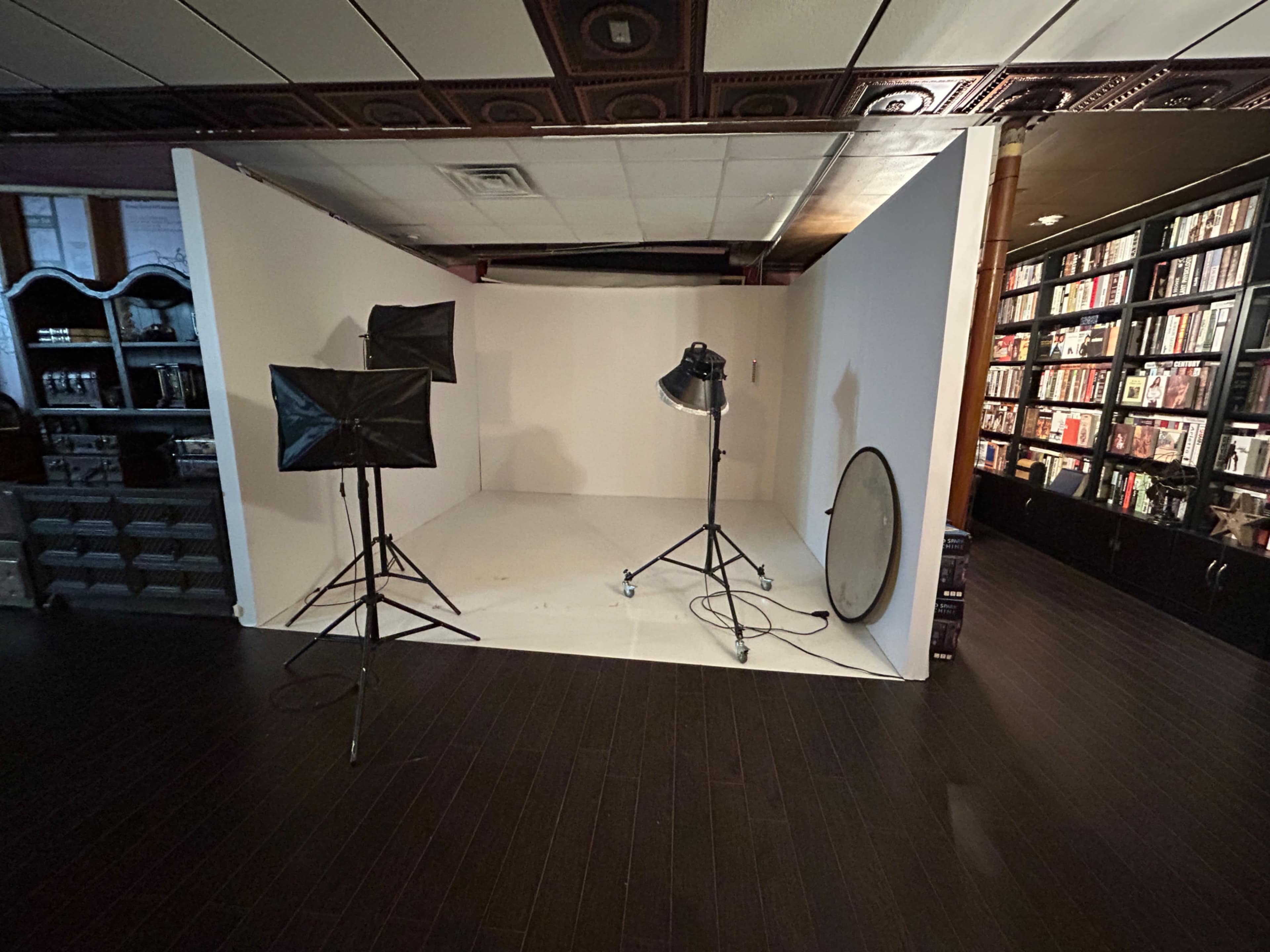The image shows a small photography studio setup with lighting equipment and a white backdrop, positioned next to a bookshelf filled with books.