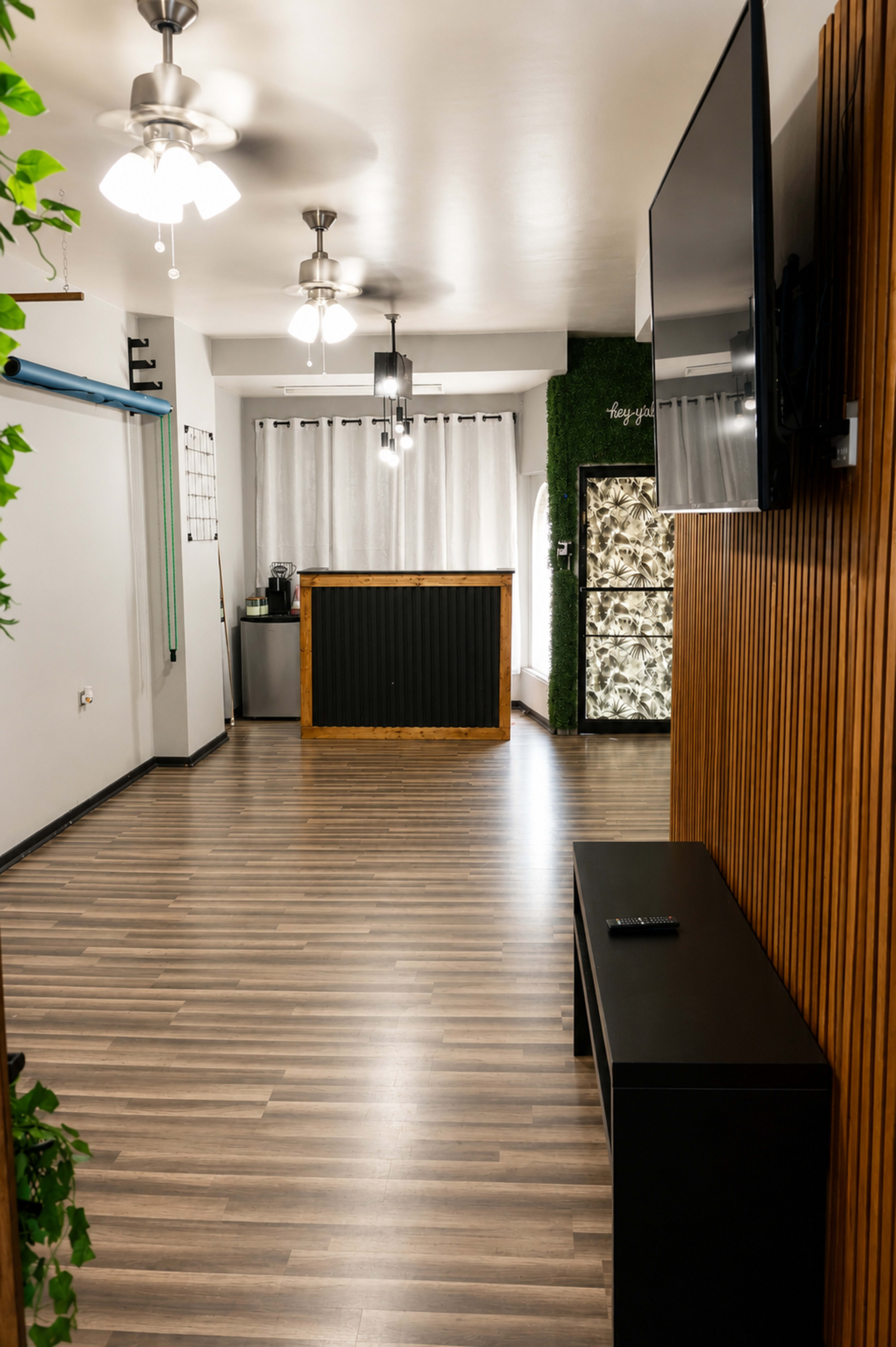 The image shows a modern, minimalist interior space with wooden flooring, a reception counter, a beverage station, and large windows covered with sheer curtains.