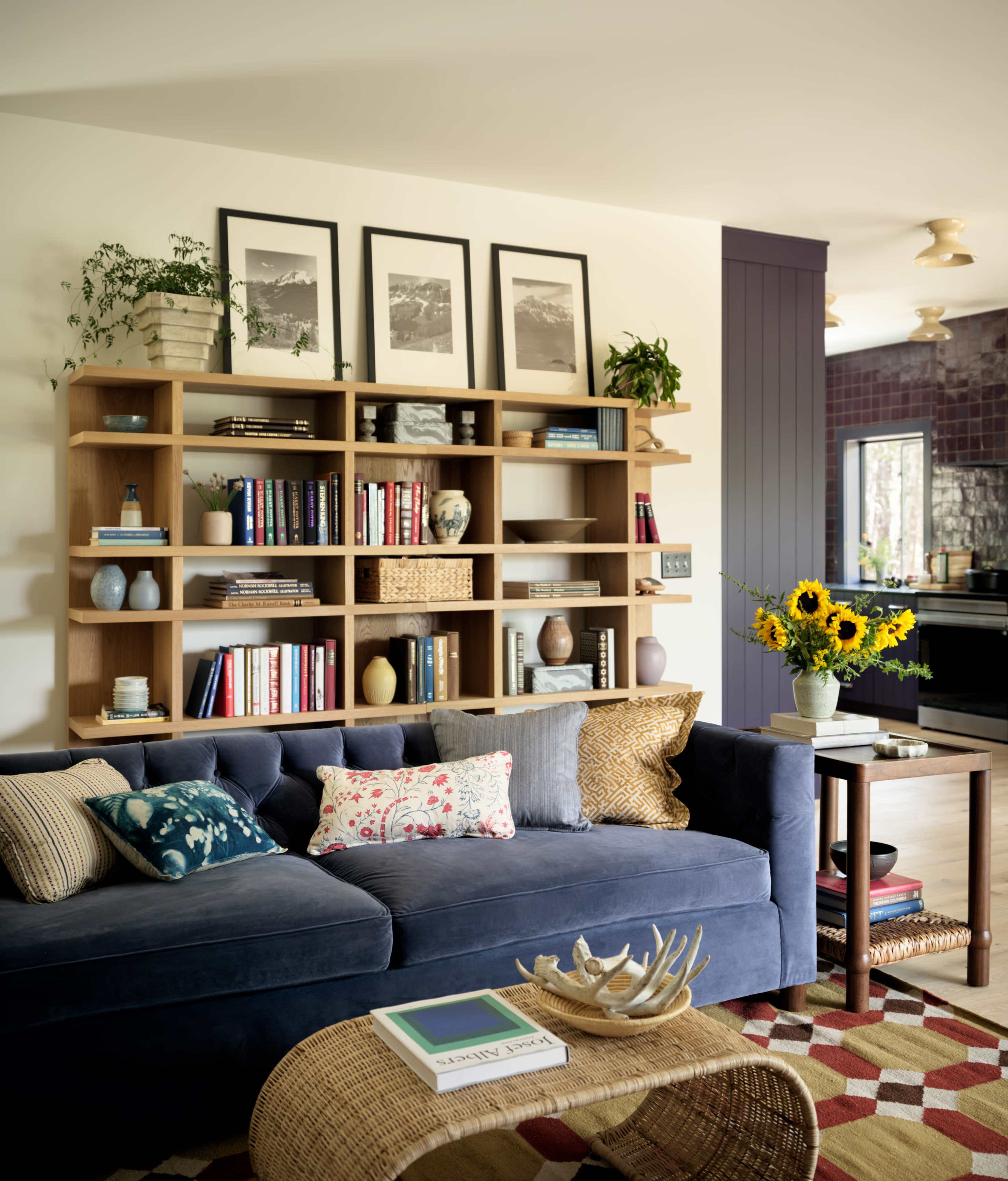 A modern living room features a blue sofa adorned with decorative pillows, a wooden bookshelf filled with books and plants, and a side table with a vase of sunflowers.