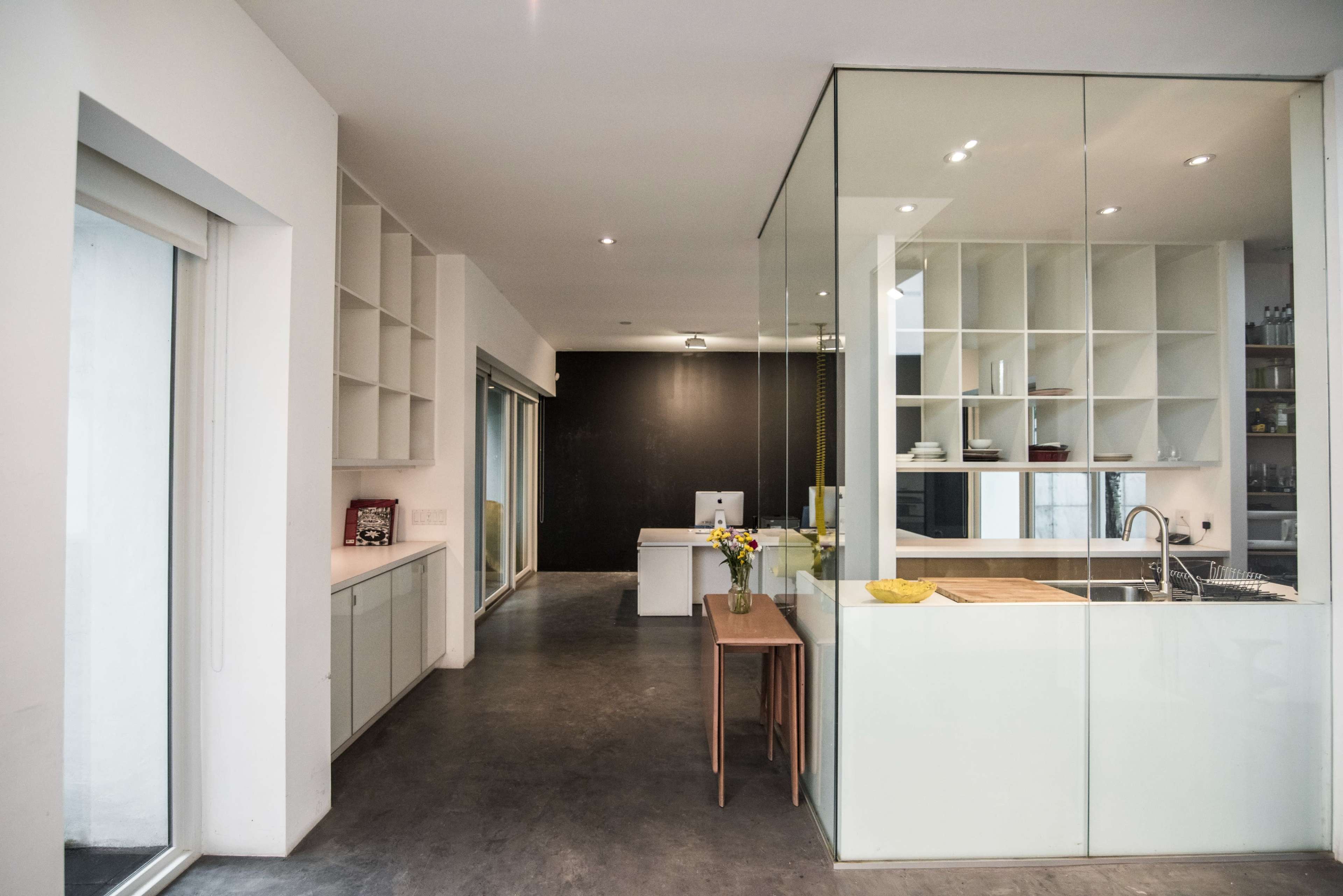 The image shows a modern kitchen and living space with glass paneling, light-colored cabinetry, and a wooden table.