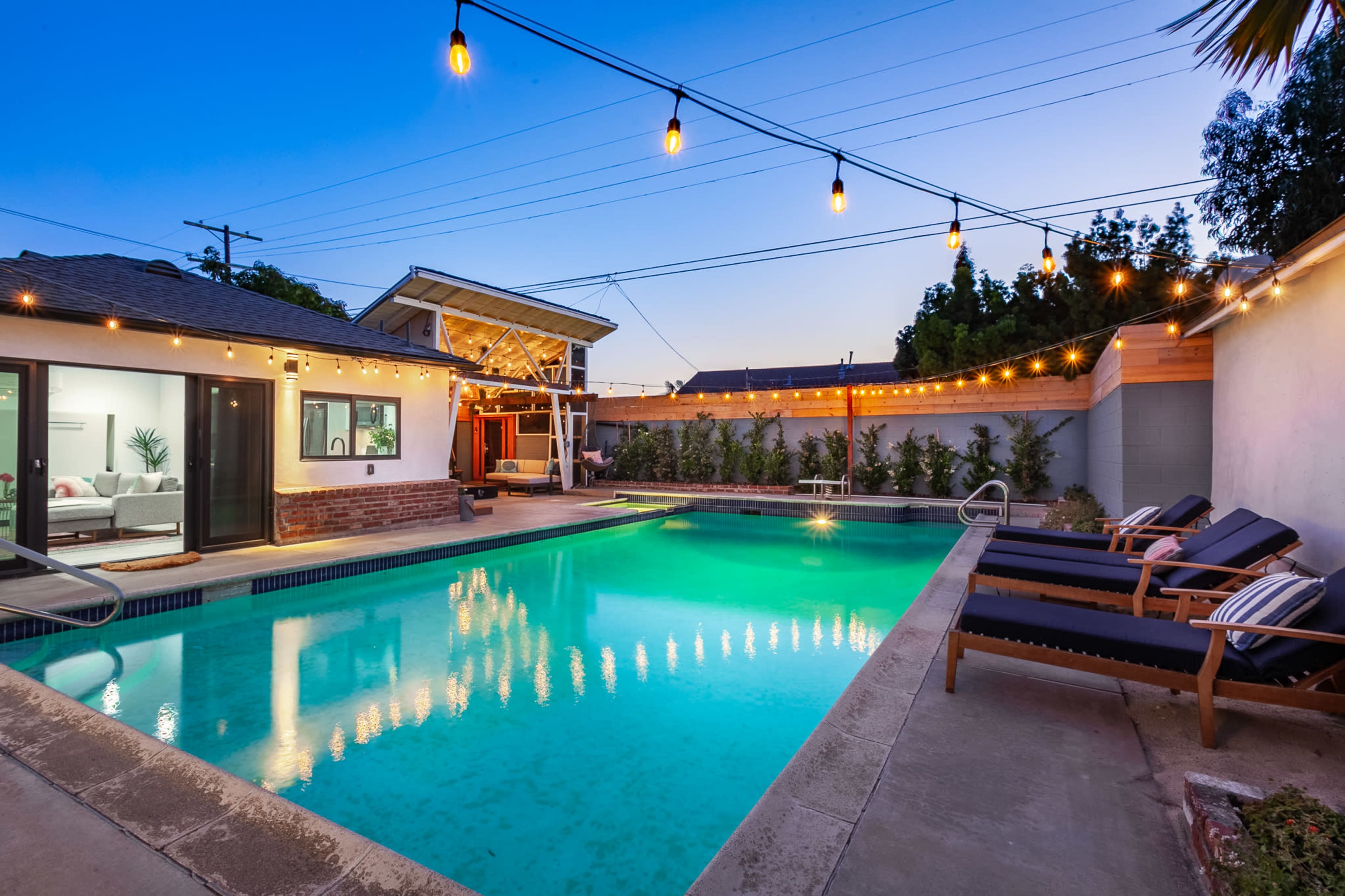 A well-lit backyard features a swimming pool surrounded by lounge chairs and string lights against a twilight sky.