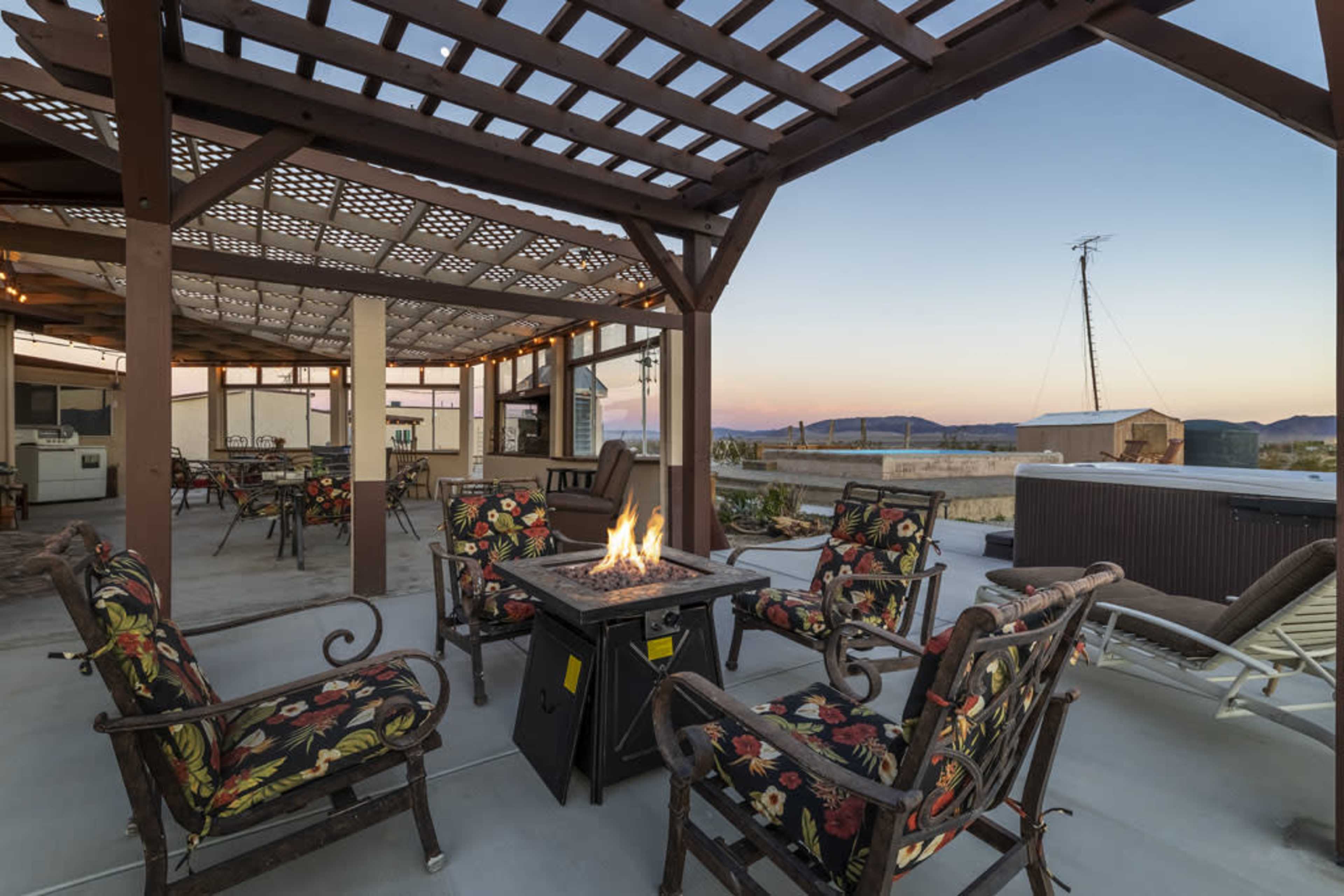 A spacious rooftop area with wooden pergolas, seating around a fire pit, and mountain views in the background during sunset.