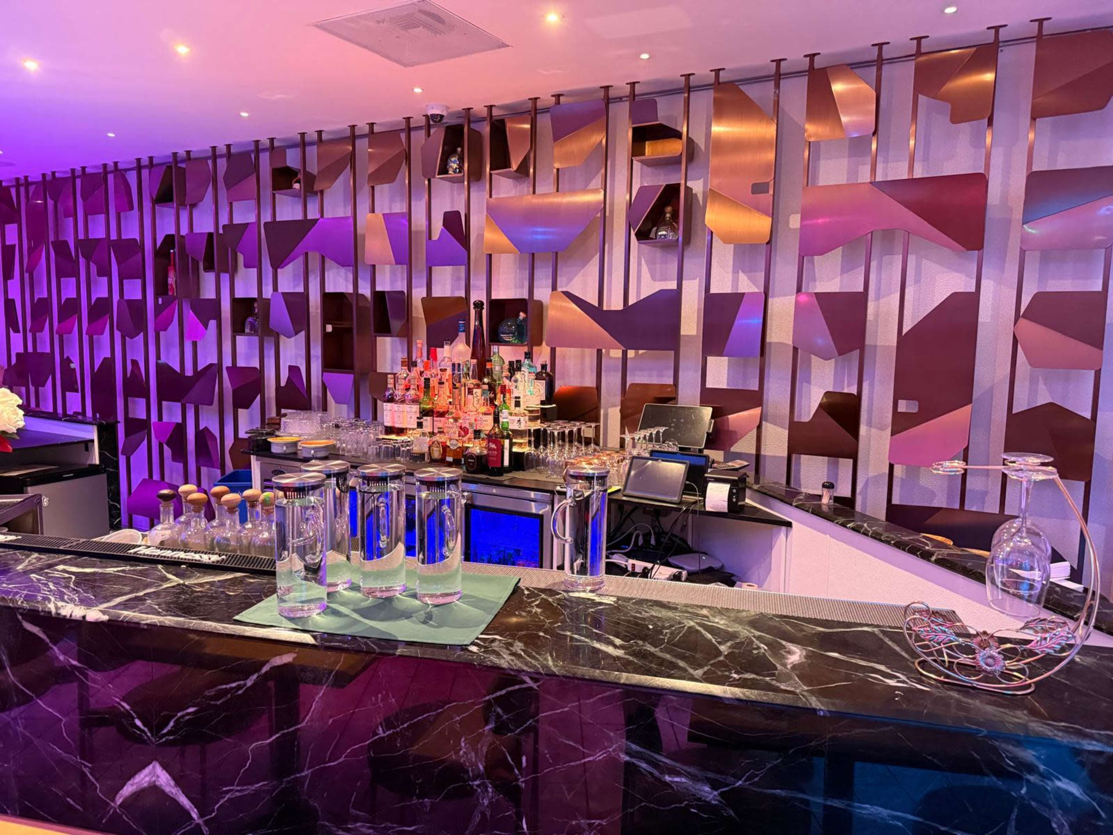The image shows a modern bar with a well-stocked shelf of liquor bottles against a stylized, colorful backdrop and a sleek countertop.