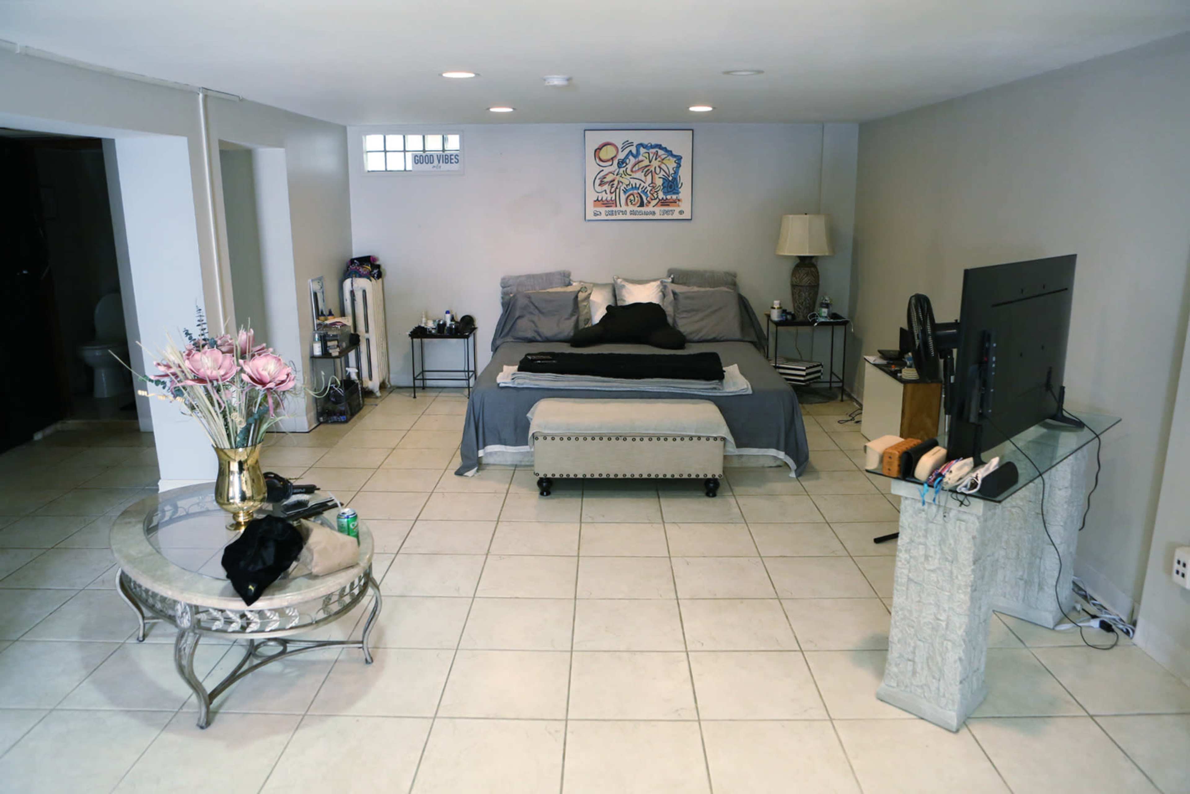 The image shows a spacious, minimalist bedroom with a bed at the center, a coffee table, and a television on a stand against a light-colored wall.