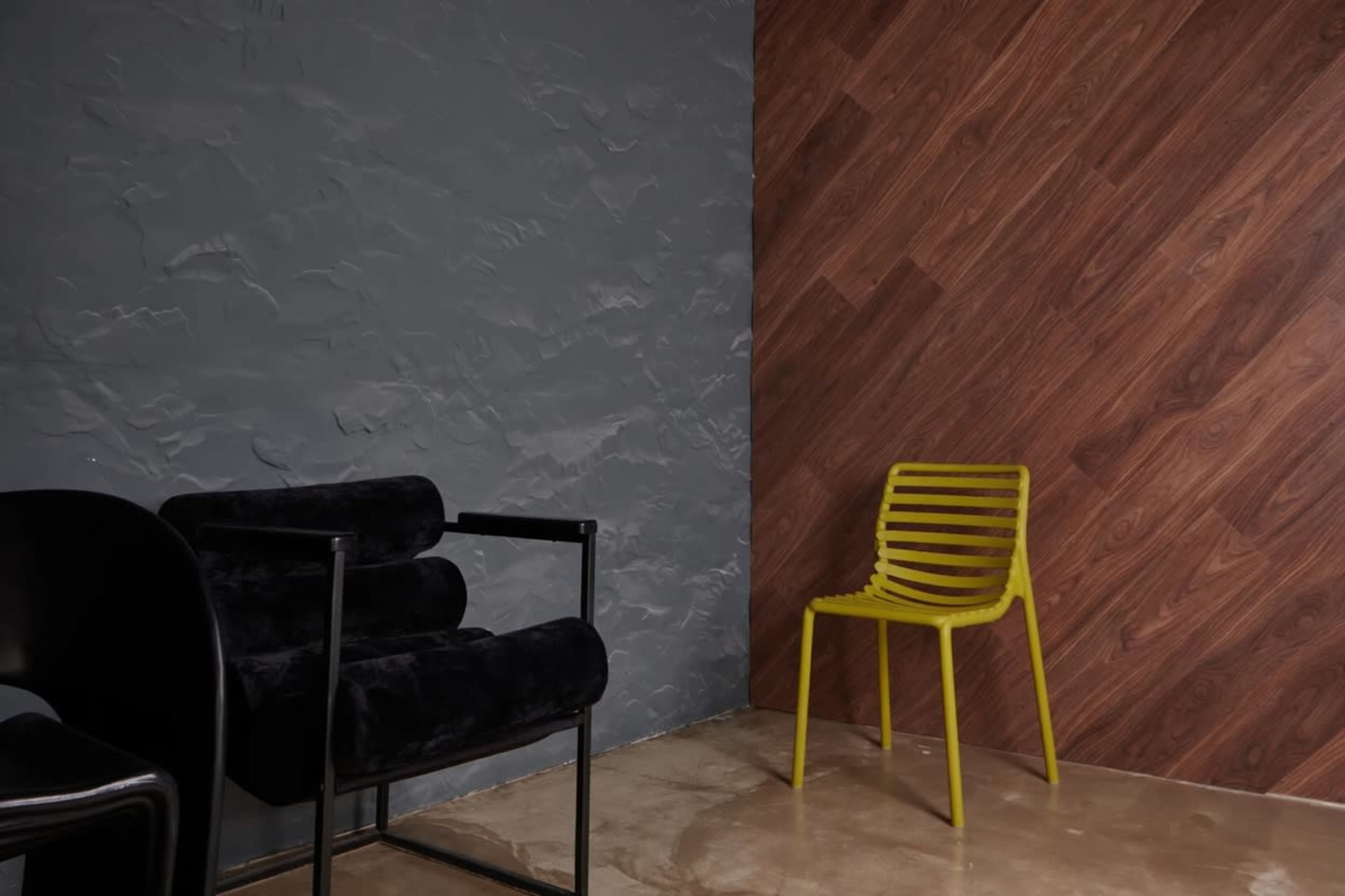 The image shows a corner of a room with a gray textured wall, a wooden wall, and two chairs: one black with a modern design and another bright yellow with horizontal slats.