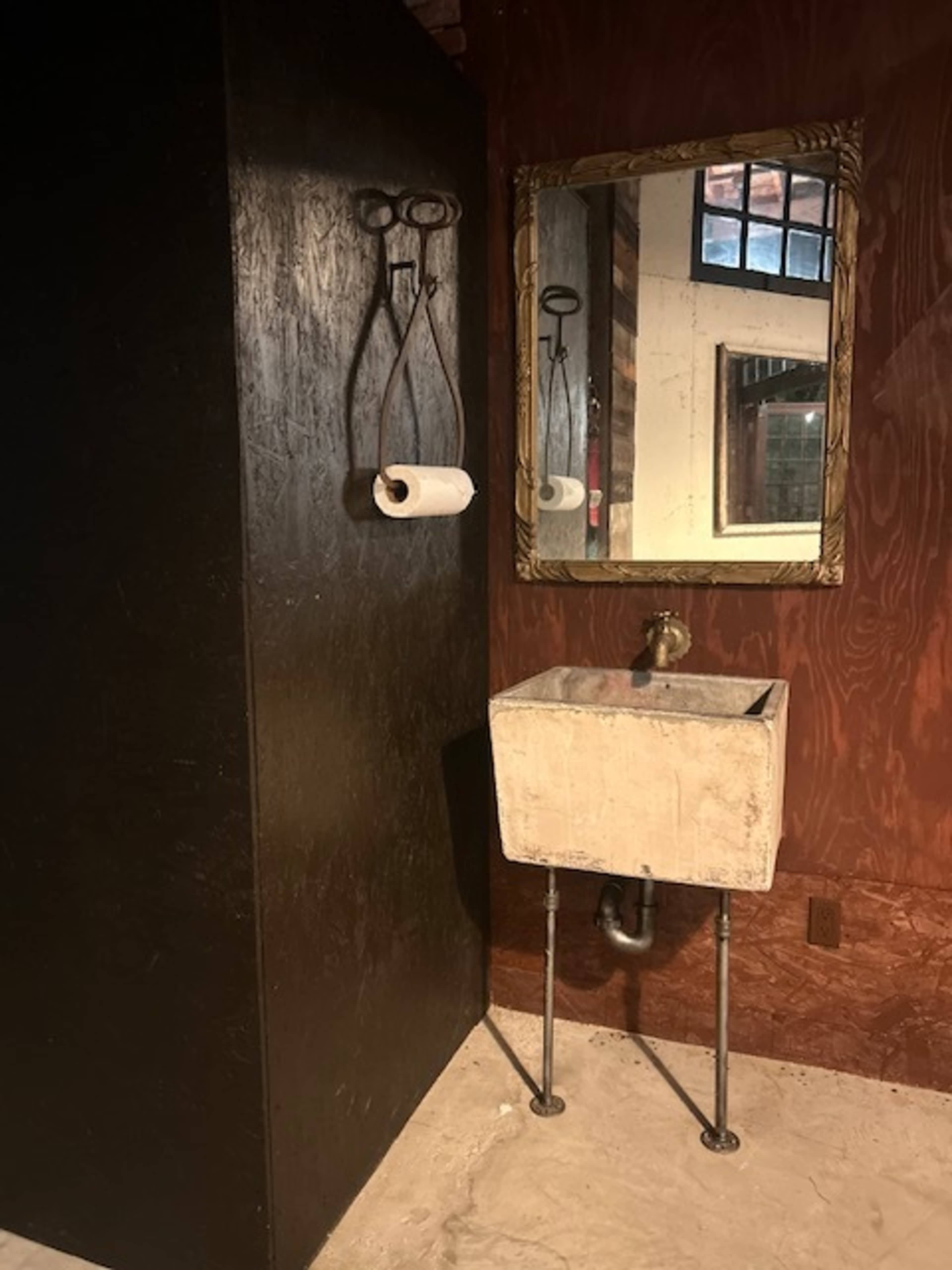 A rustic bathroom with a black cabinet, a white sink on metal legs, and a framed mirror reflected in the background.