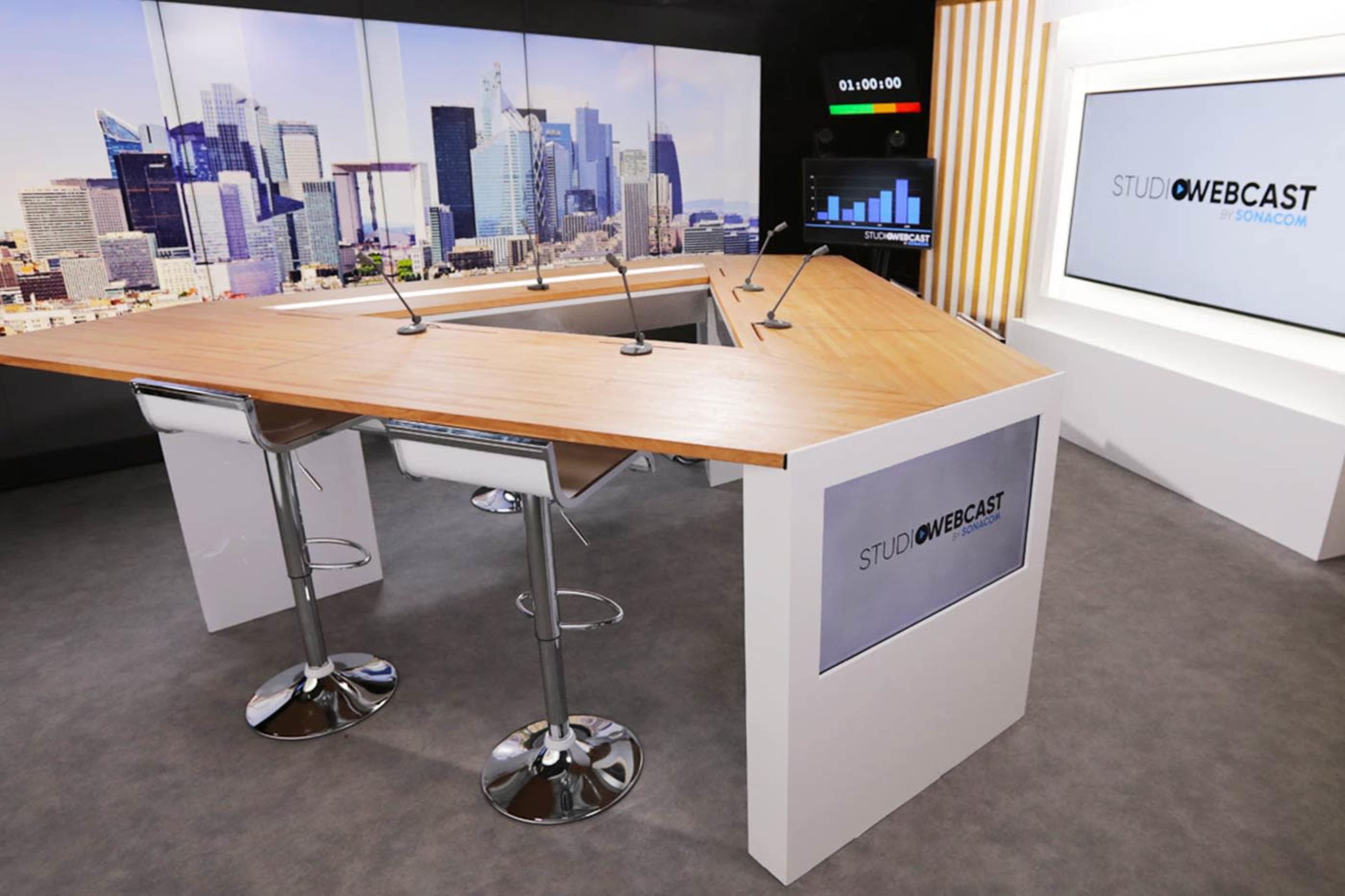 A modern broadcast studio with a segmented desk and tall bar stools, set against a backdrop of a city skyline.