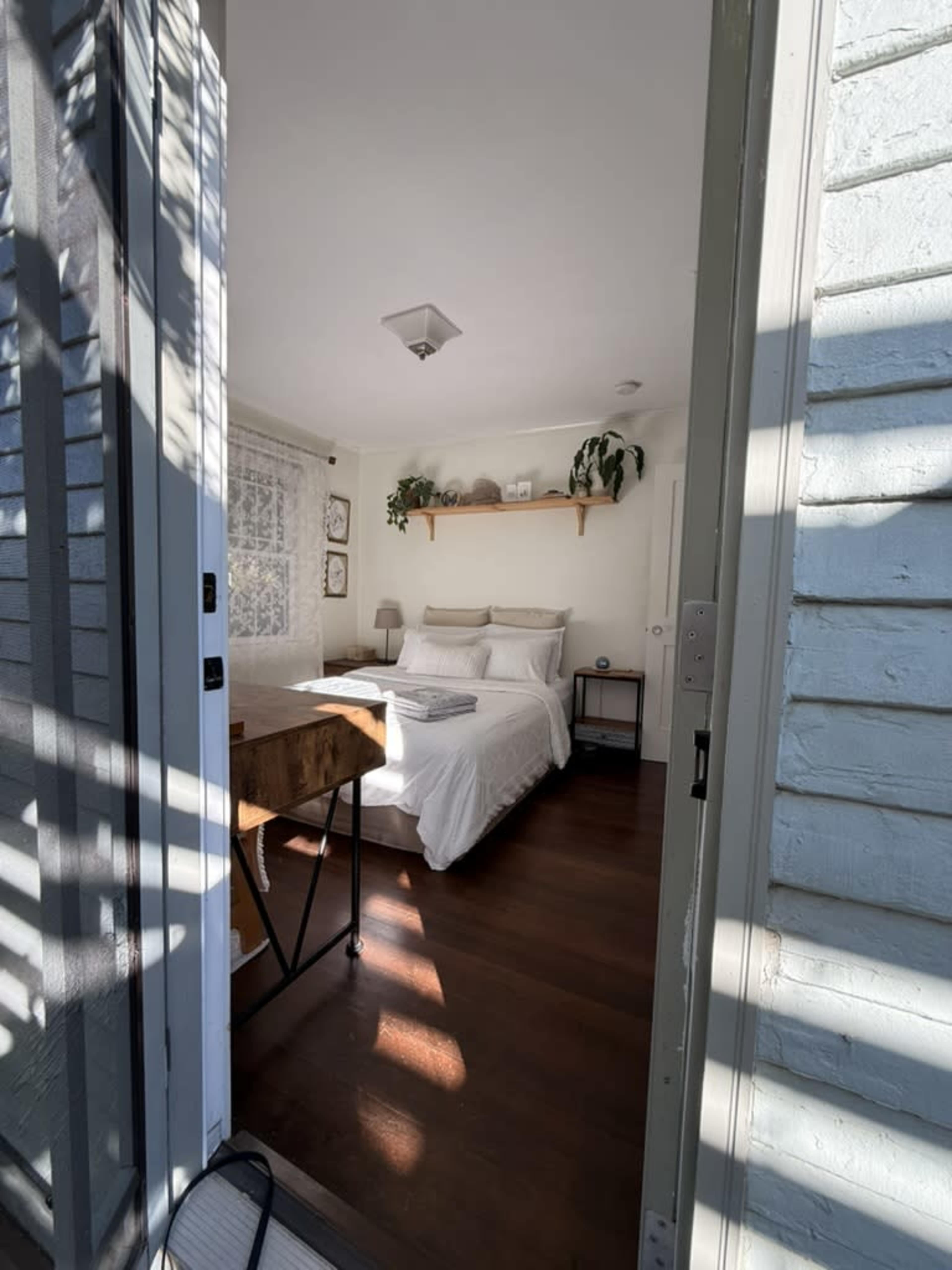 A bright bedroom with a white bed, wooden floor, and a shelf with plants is visible through an open door.