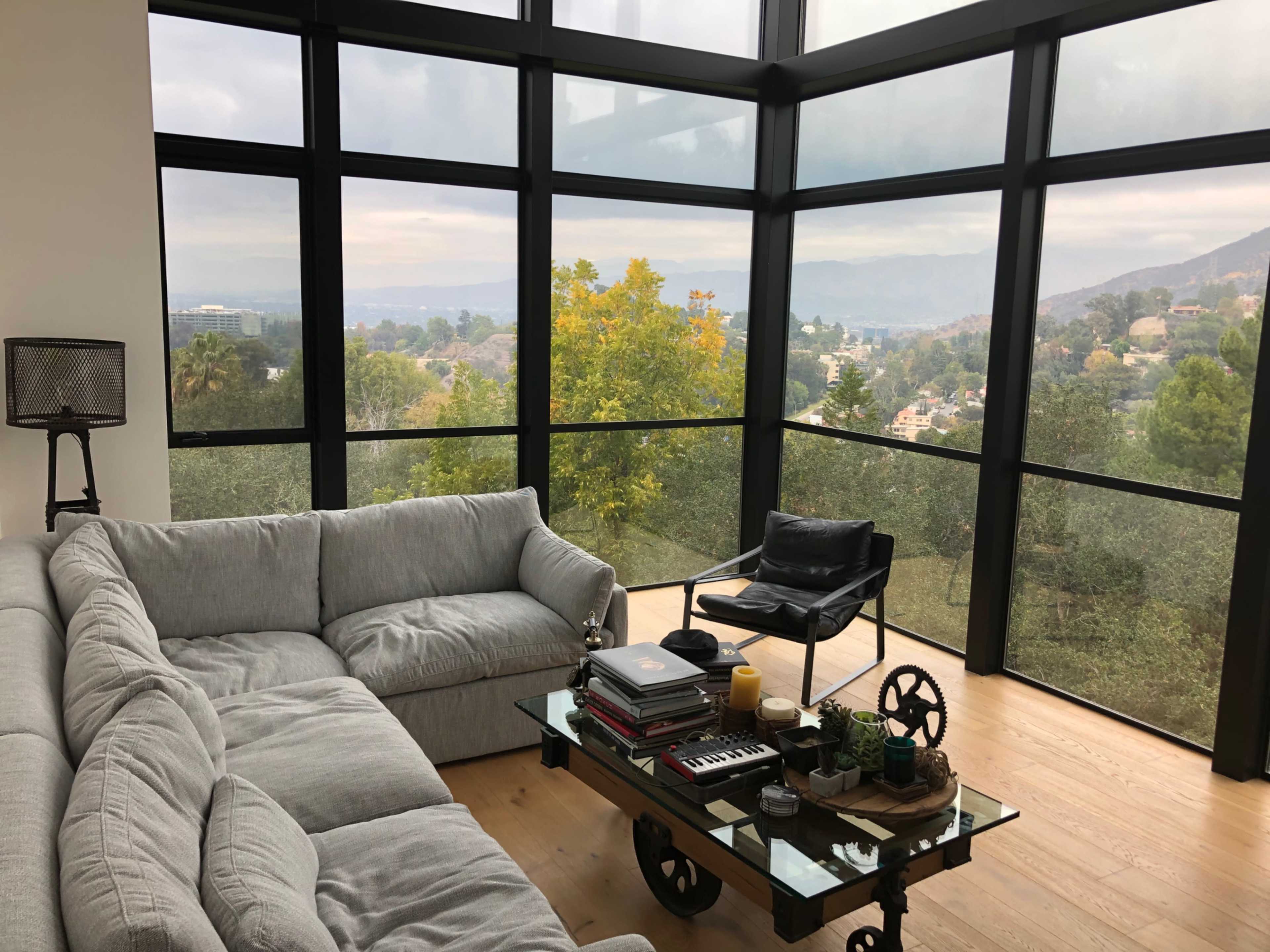 A modern living room features a sectional couch and a coffee table with various items, surrounded by large windows that offer a view of trees and hills outside.