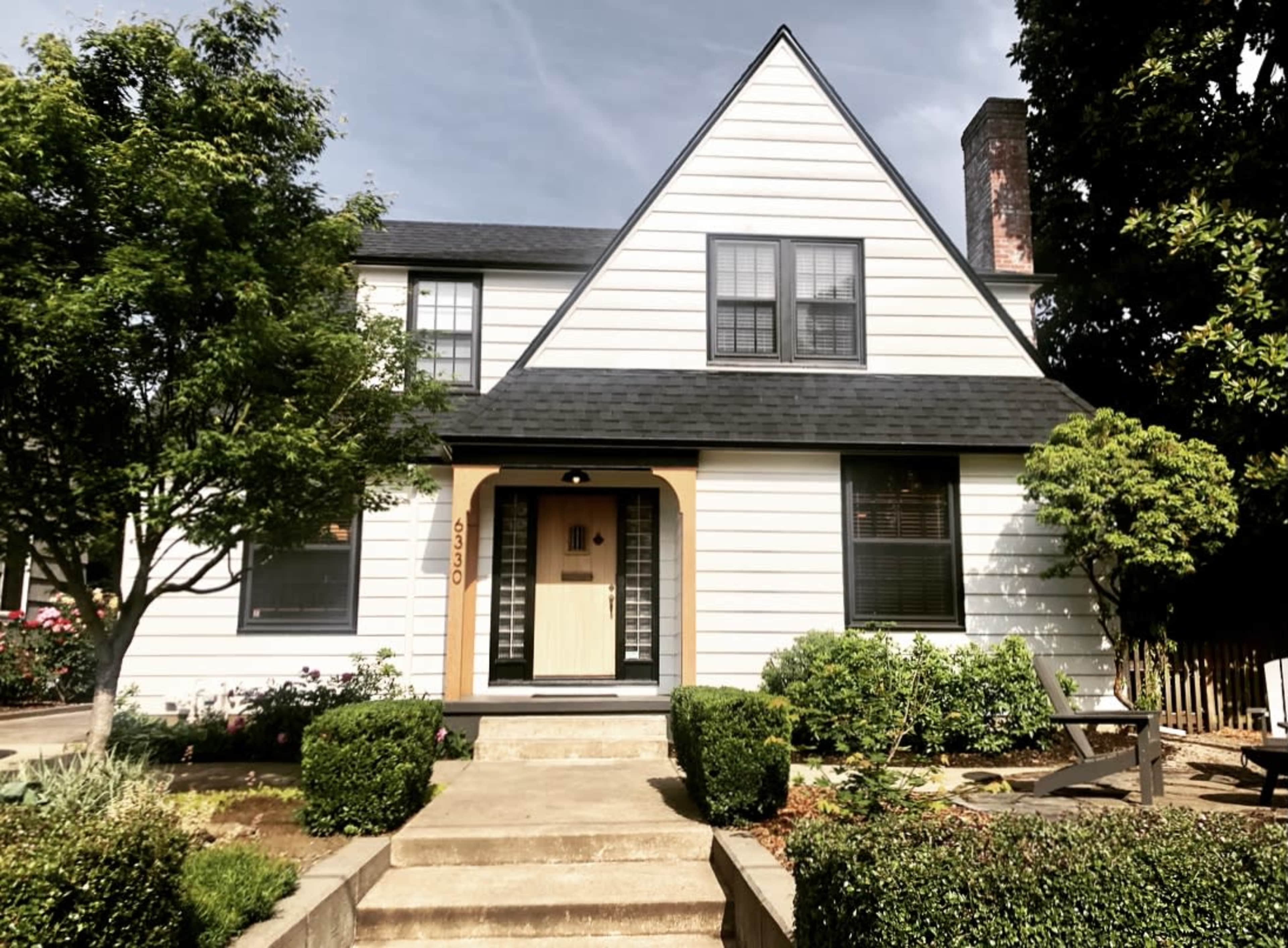 A two-story, A-frame style house with a white exterior, black roof, and a landscaped front yard.