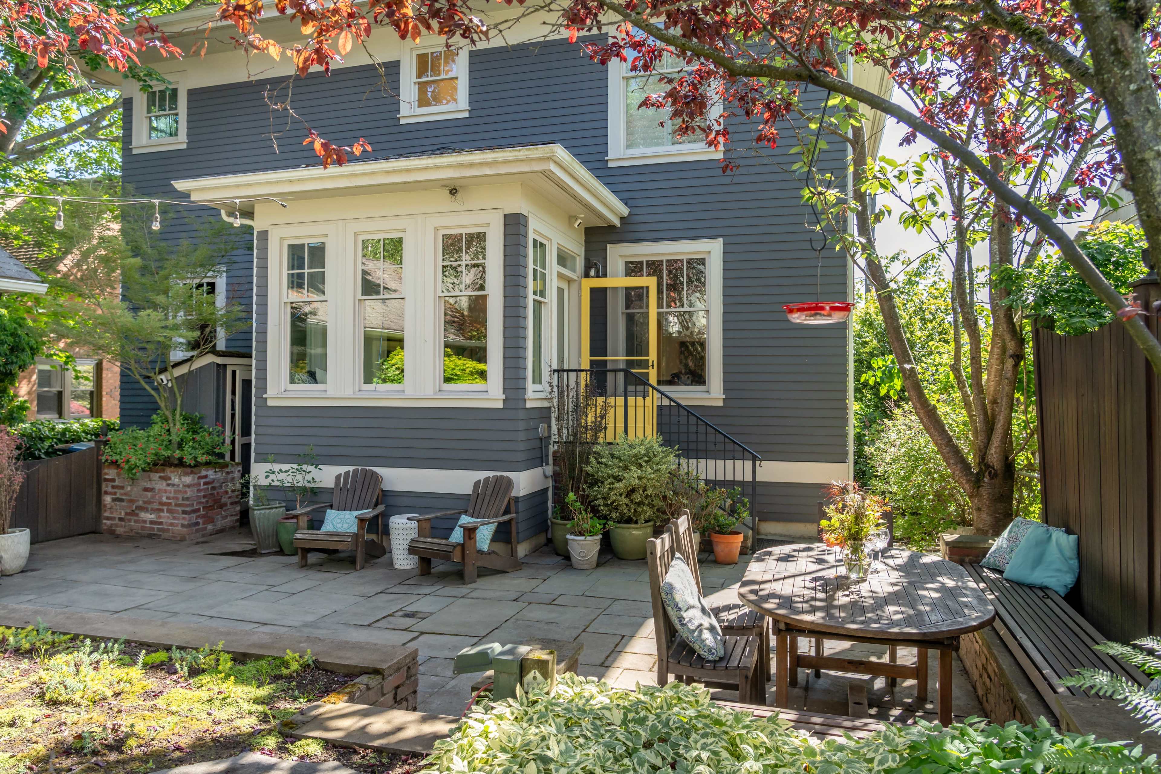 A patio area with wooden furniture and a dining table is situated next to a house featuring large windows and a garden with various plants.