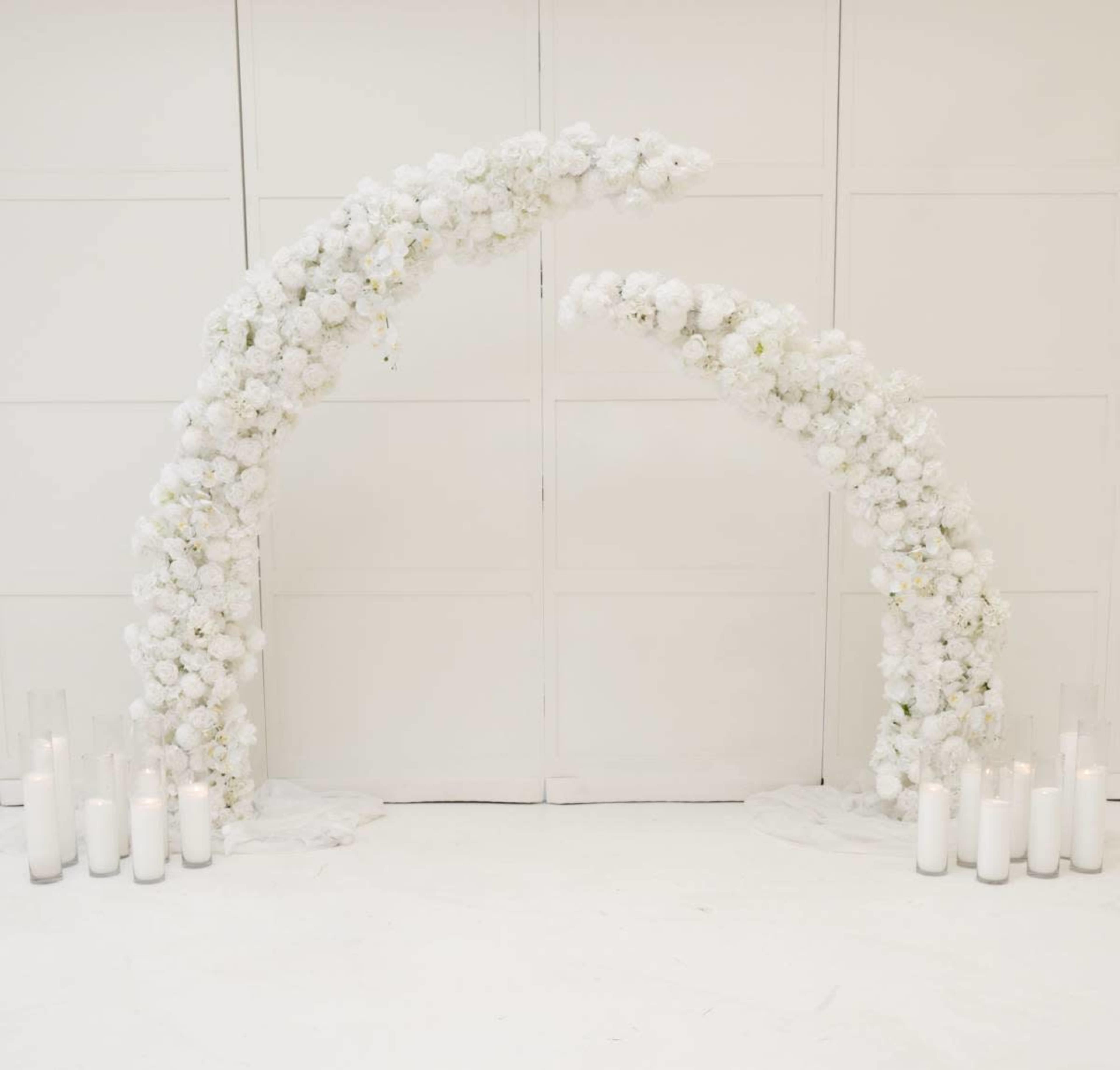 The image shows an elegant floral arch made of white flowers with candles arranged in glass holders on either side.