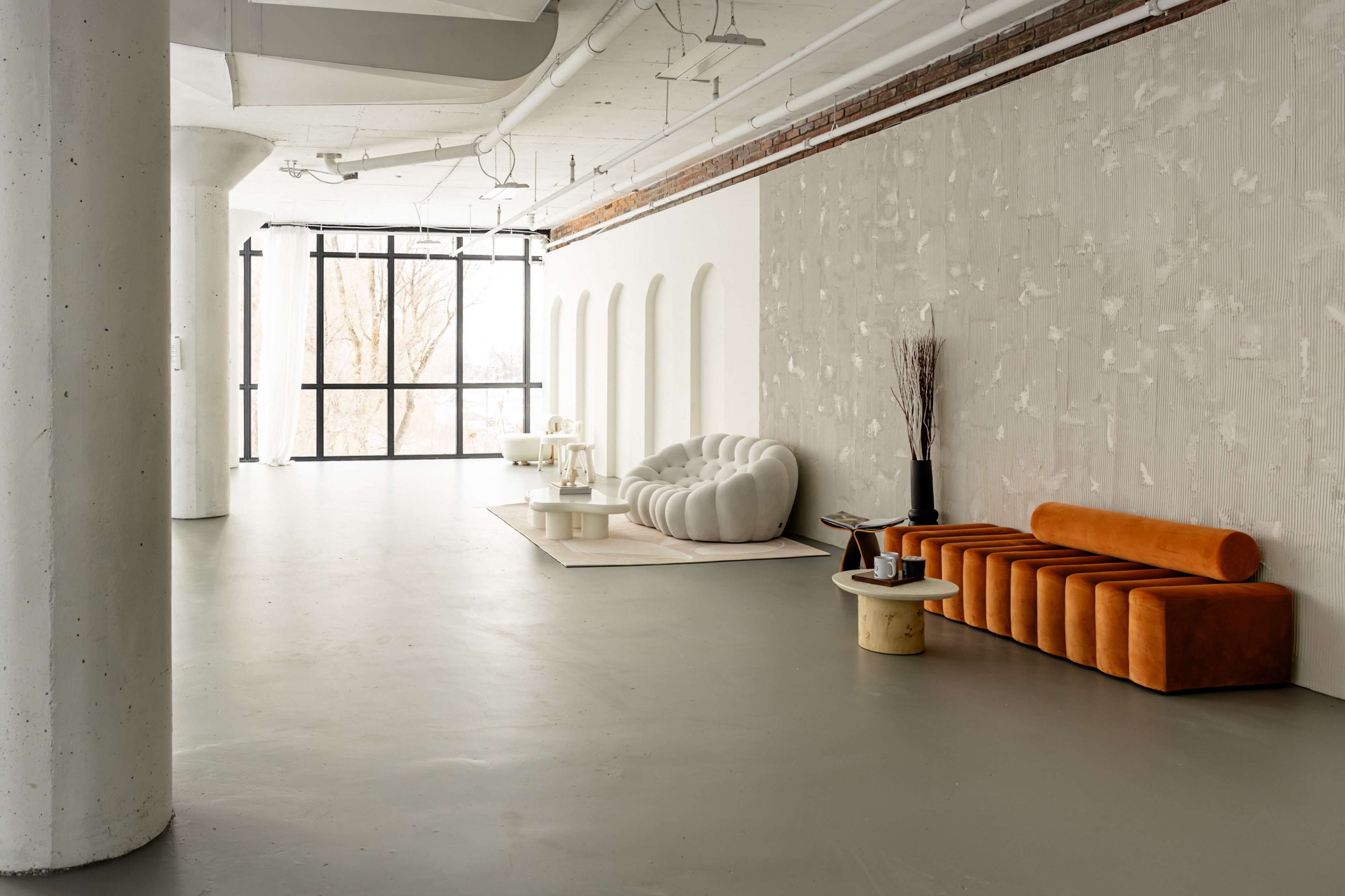 The image features a spacious interior with minimalist furniture, including a white textured wall, an orange upholstery sofa, and contemporary seating arrangements.