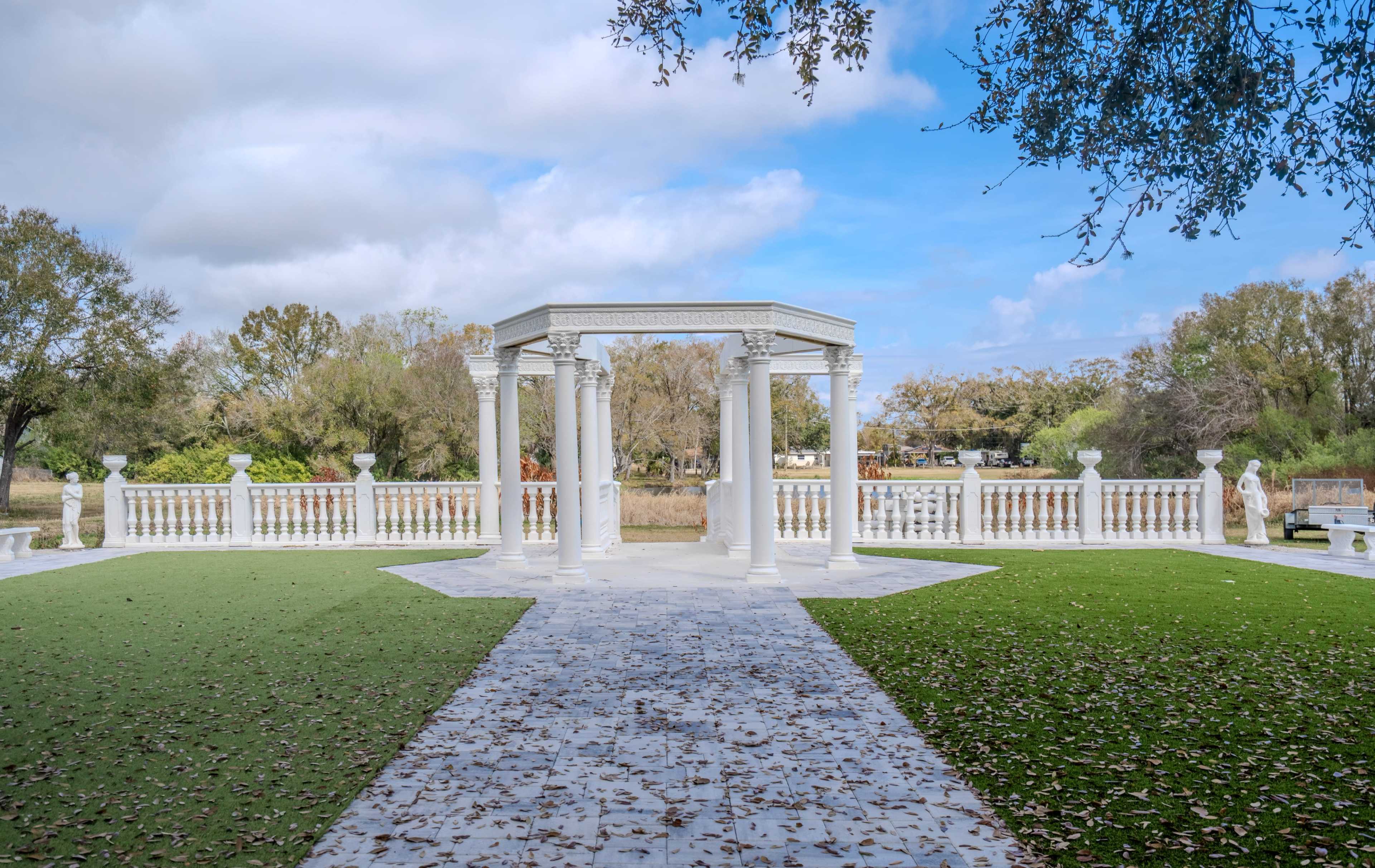 A white gazebo with columns stands on a grassy area surrounded by trees and a path leading to a body of water in the background.