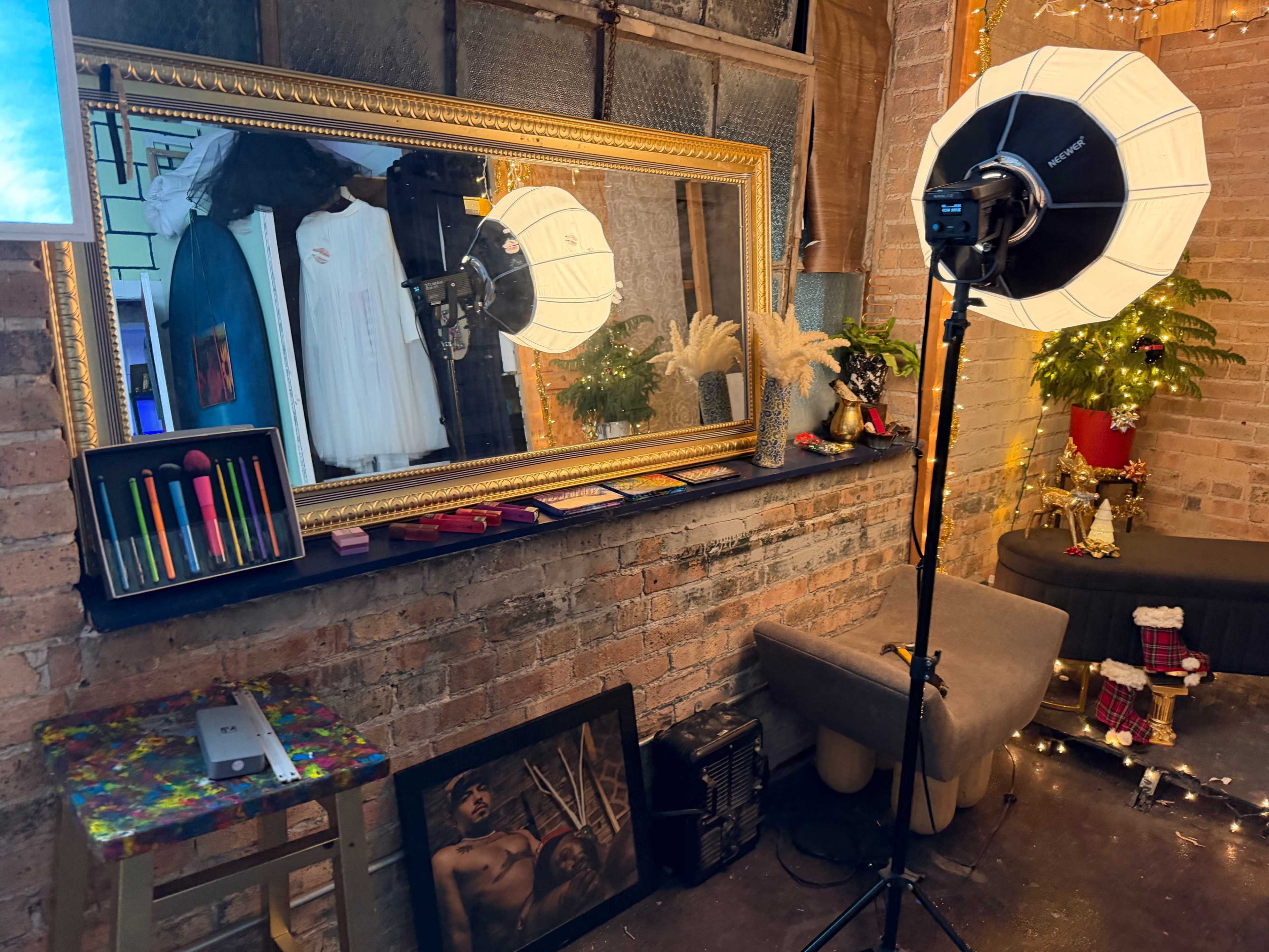 The image shows a styled interior space featuring a large ornate mirror, studio lighting equipment, a decorative table with art supplies, and a plush seating area adorned with plants and festive lighting.
