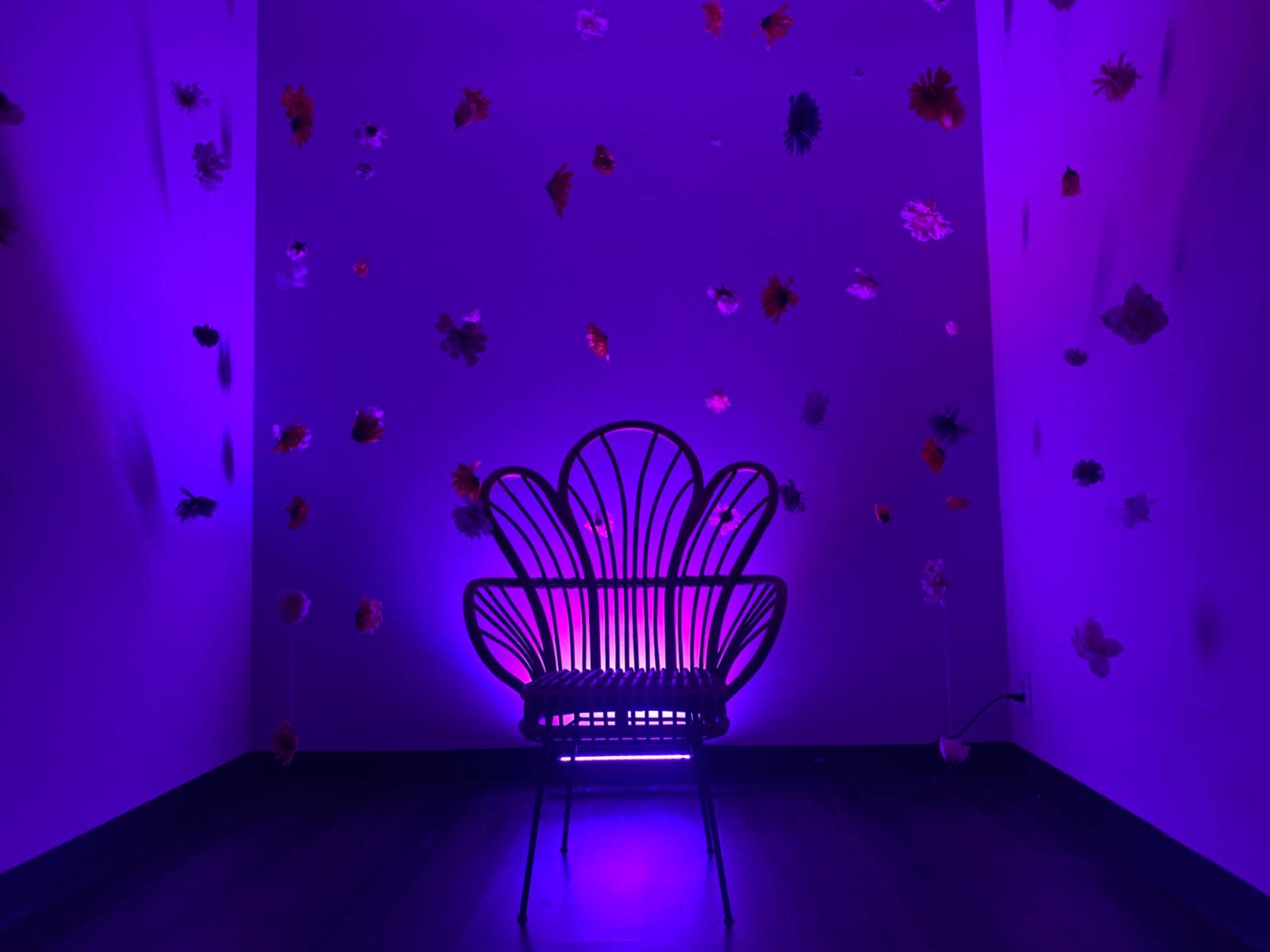 A shell-shaped chair is illuminated by purple light in a room adorned with colorful artificial flowers hanging from the walls.